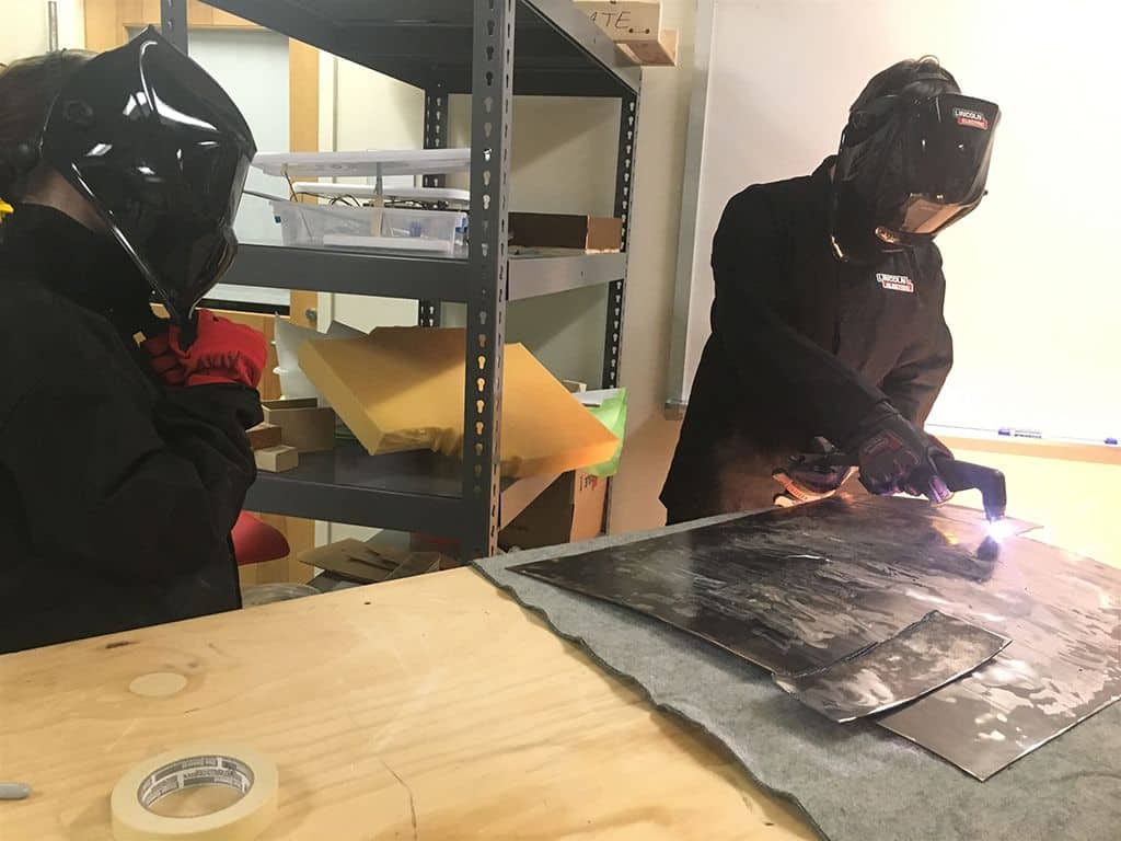 Two individuals in black jackets collaborate on shaping a piece of metal in a workshop setting.