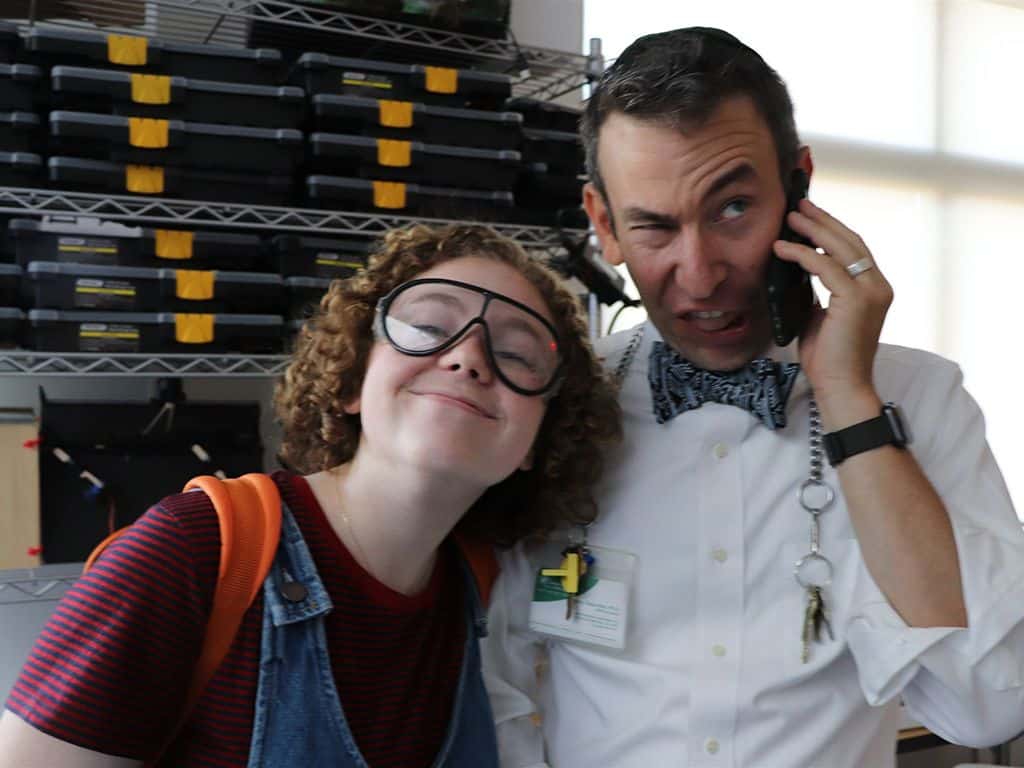 A teacher and a student standing side by side, both smiling and dressed casually.