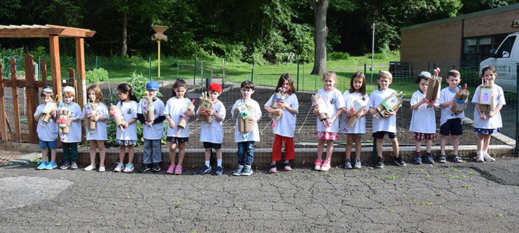 Children in white shirts stand in a line outdoors, each holding a crafted creation. The backdrop is a garden with lush greenery and a building. The mood is cheerful and playful.
