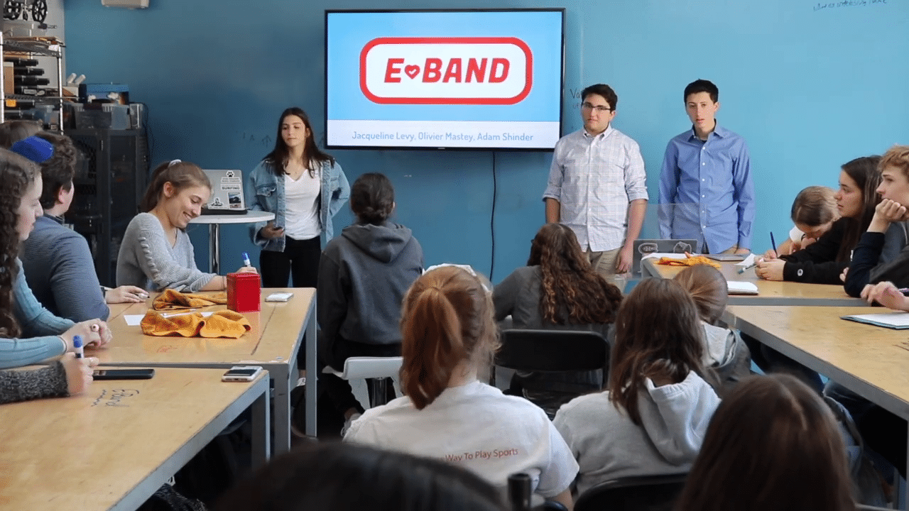 A group of people seated at tables, attentively watching a presentation on a large screen in front of them.