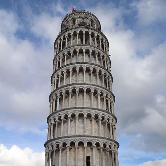 The Leaning Tower of Pisa set against a partly cloudy blue sky, showcasing its distinct tilt and intricate architecture. A red flag waves atop the tower.