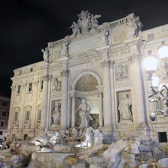 Illuminated Trevi Fountain at night in Rome, featuring ornate sculptures and a grand facade with classical columns. A tranquil, majestic ambiance.