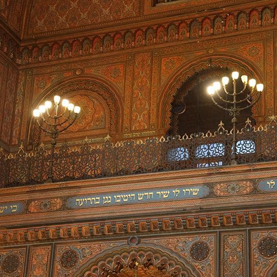 Ornate interior with intricate patterns and arches, featuring two large chandeliers casting a warm glow. Hebrew script adorns a decorative panel.