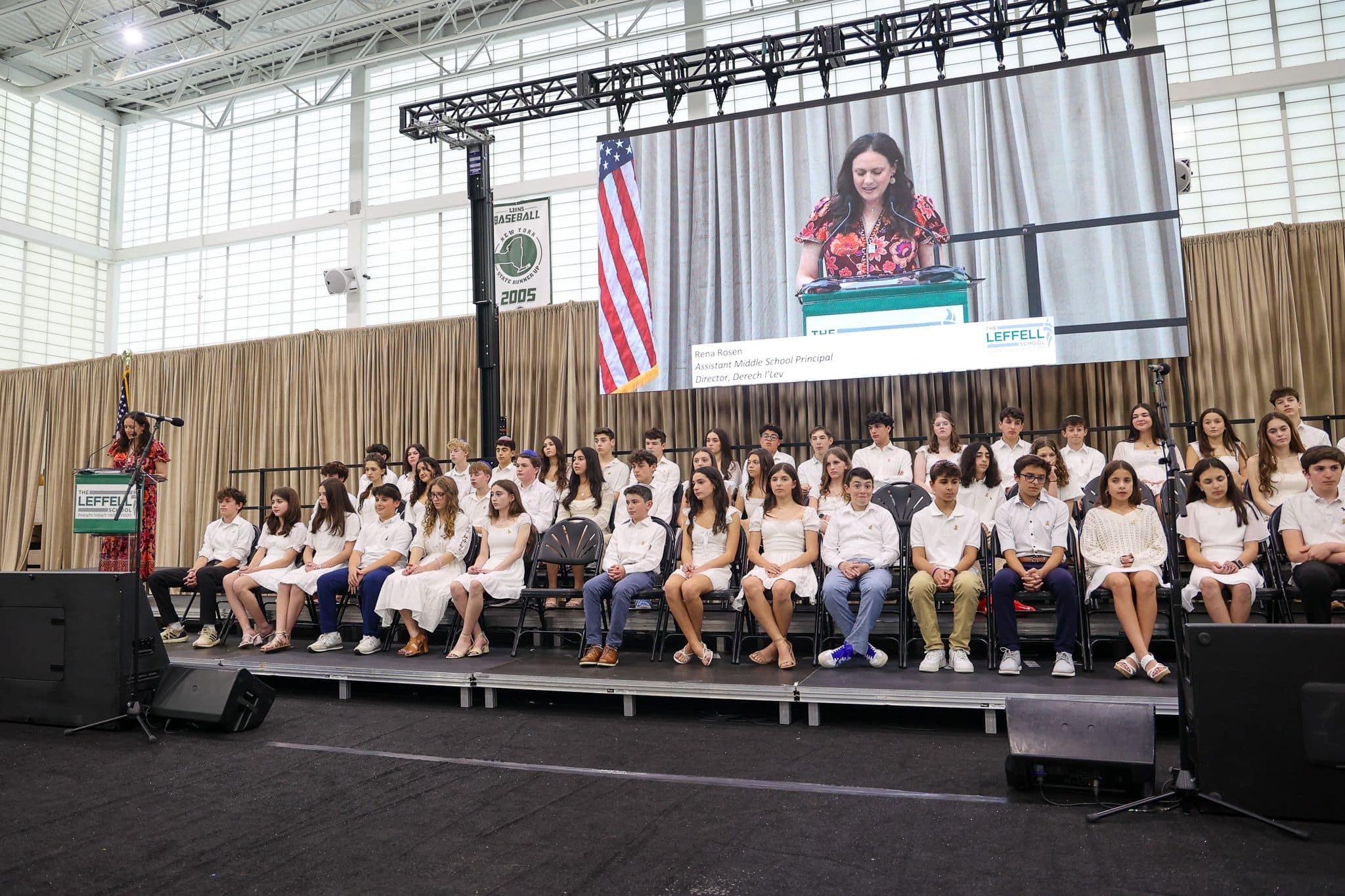A group of students in formal attire sit on stage at a school event. A woman speaks at a podium, projected on a large screen, under a U.S. flag. The setting is formal and celebratory.
