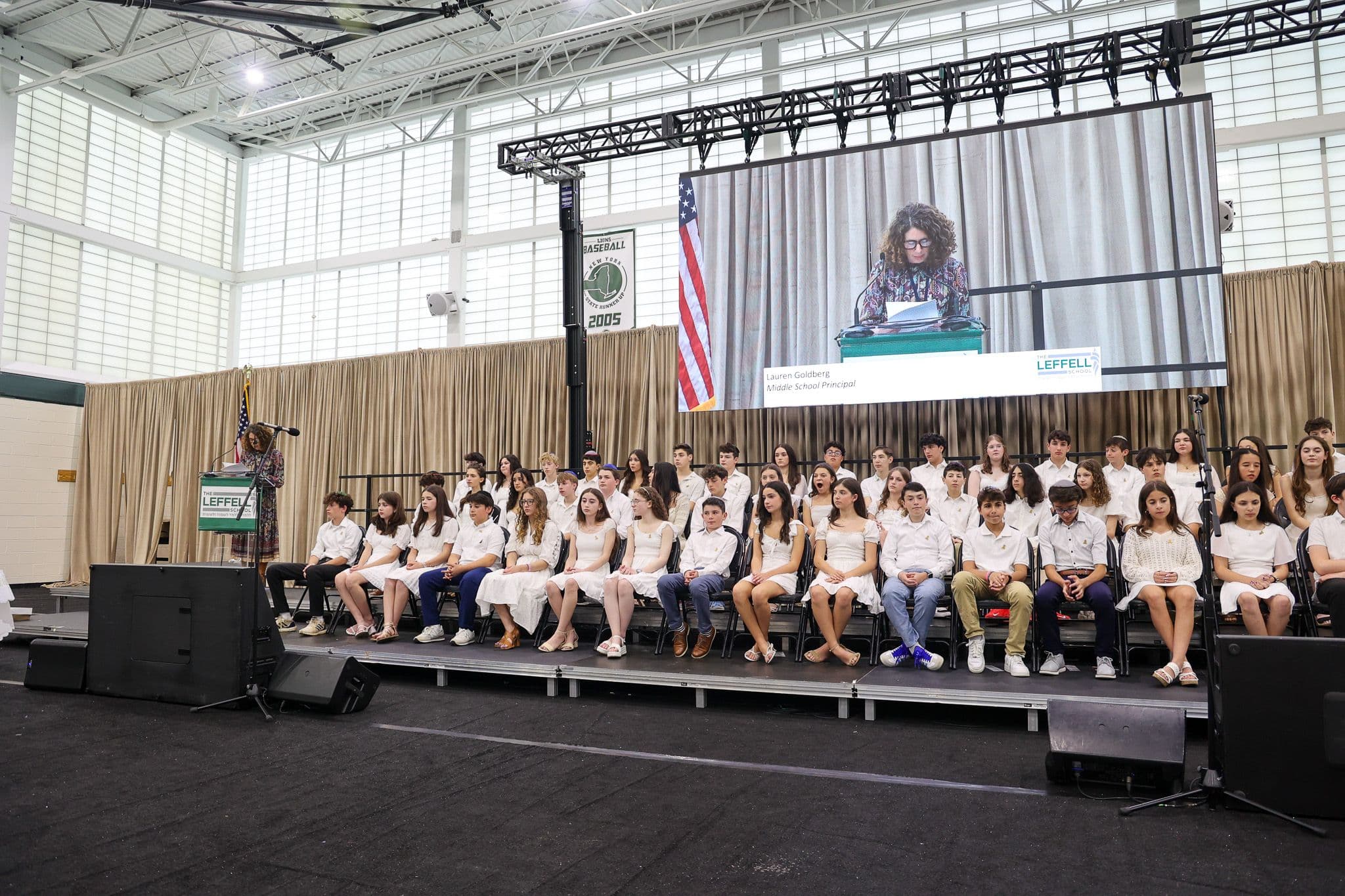 A group of students in formal attire sit on stage at a school event. A woman speaks at a podium, projected on a large screen, under a U.S. flag. The setting is formal and celebratory.