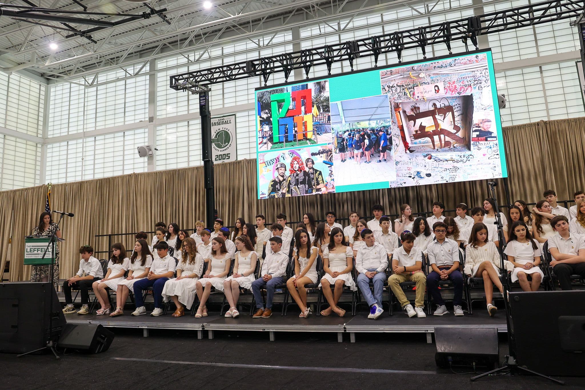 A group of students in formal attire sit on stage at a school event. A woman speaks at a podium, projected on a large screen, under a U.S. flag. The setting is formal and celebratory.