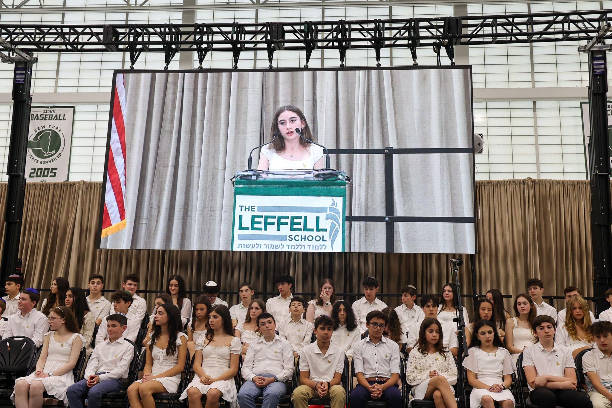 A young person speaks at a podium labeled "The Leffell School" on a large screen. Seated in front, students in white attire listen attentively. The setting is academic and formal, with a backdrop of stage curtains and an American flag.