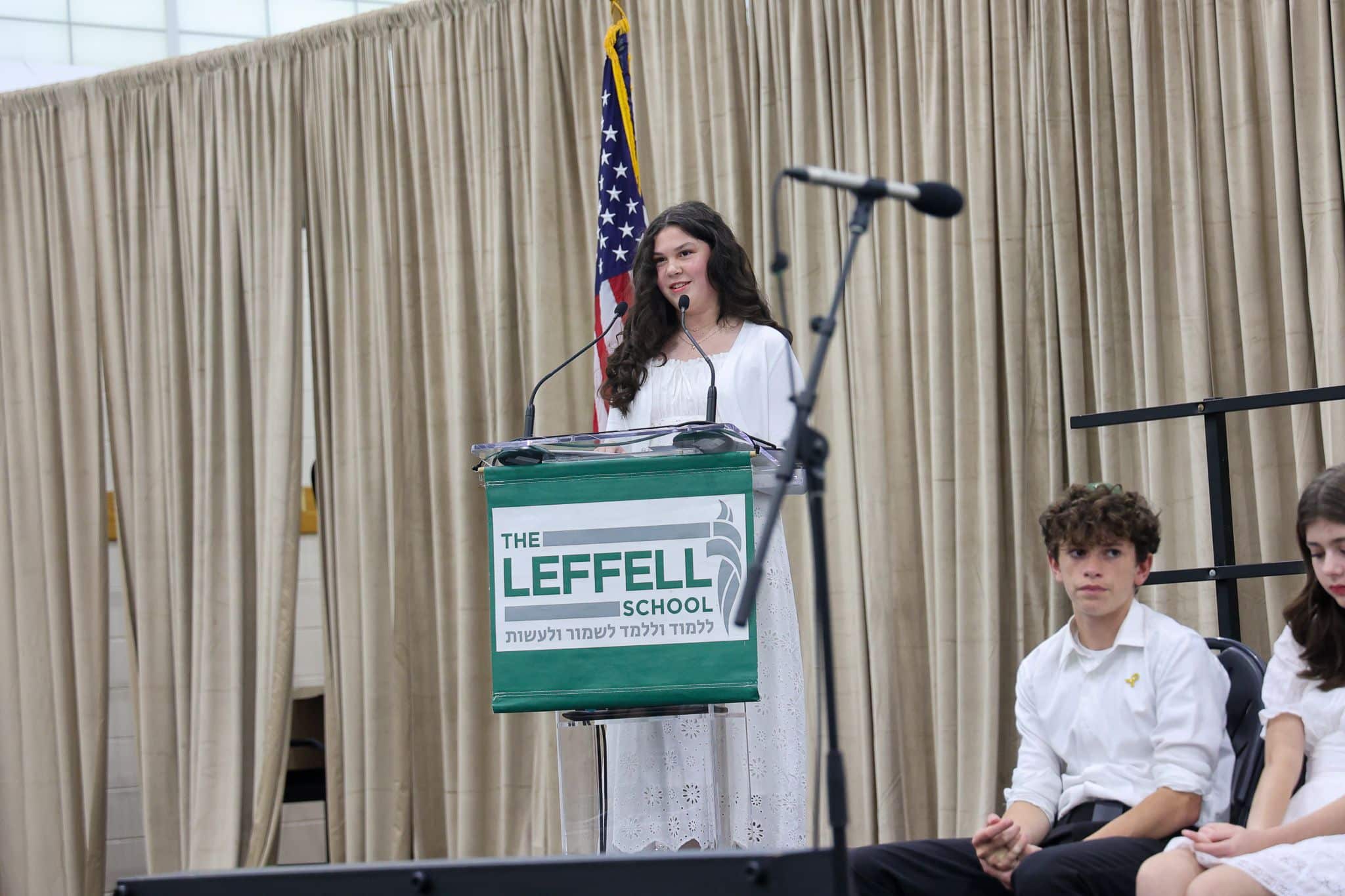 A young person speaks at a podium labeled "The Leffell School," with an American flag on the left. They wear a white outfit, set against beige curtains.