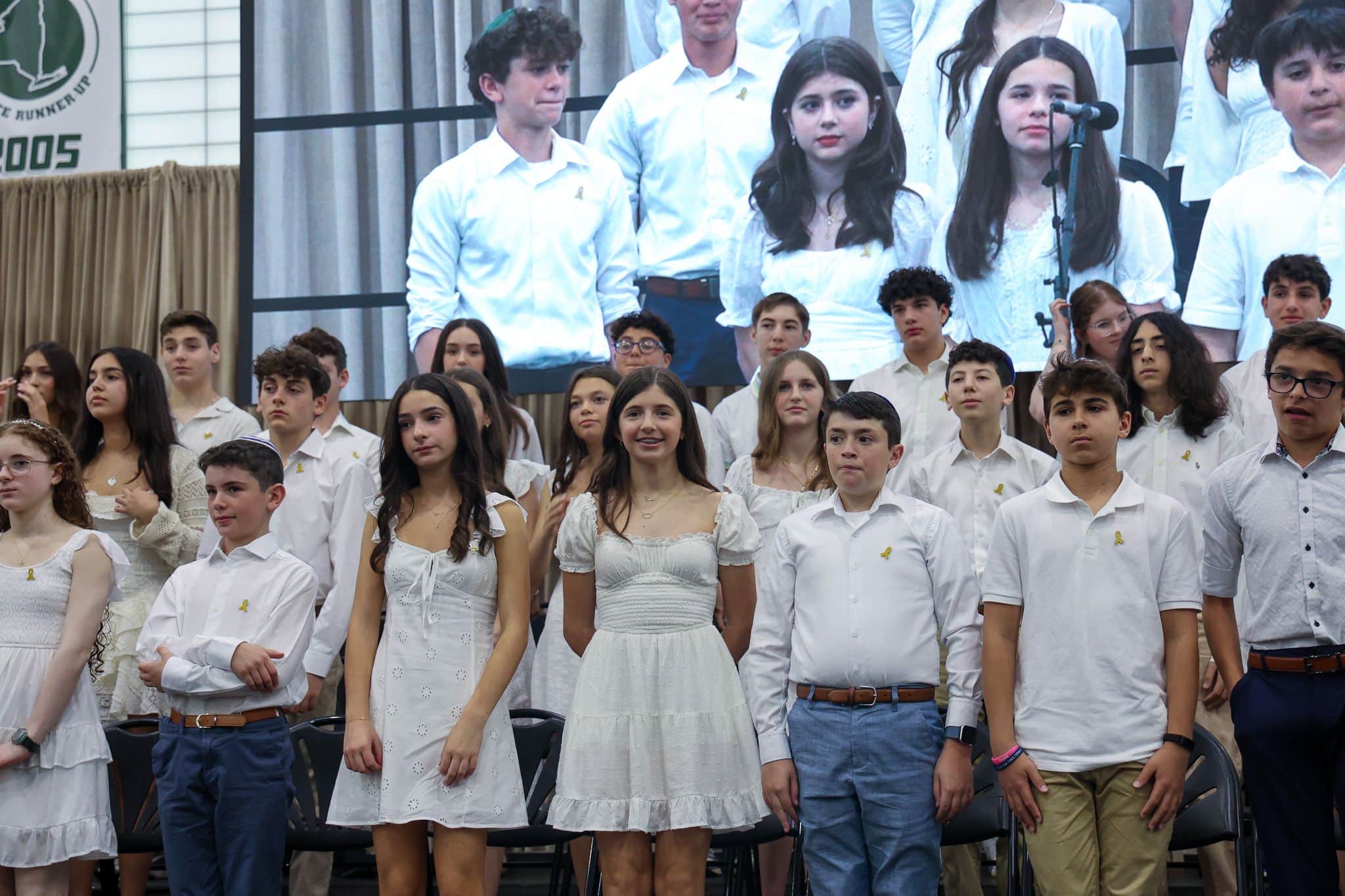 A diverse group of students stands on a stage, wearing white outfits. A large screen behind shows a close-up of two students. The mood is formal and celebratory.