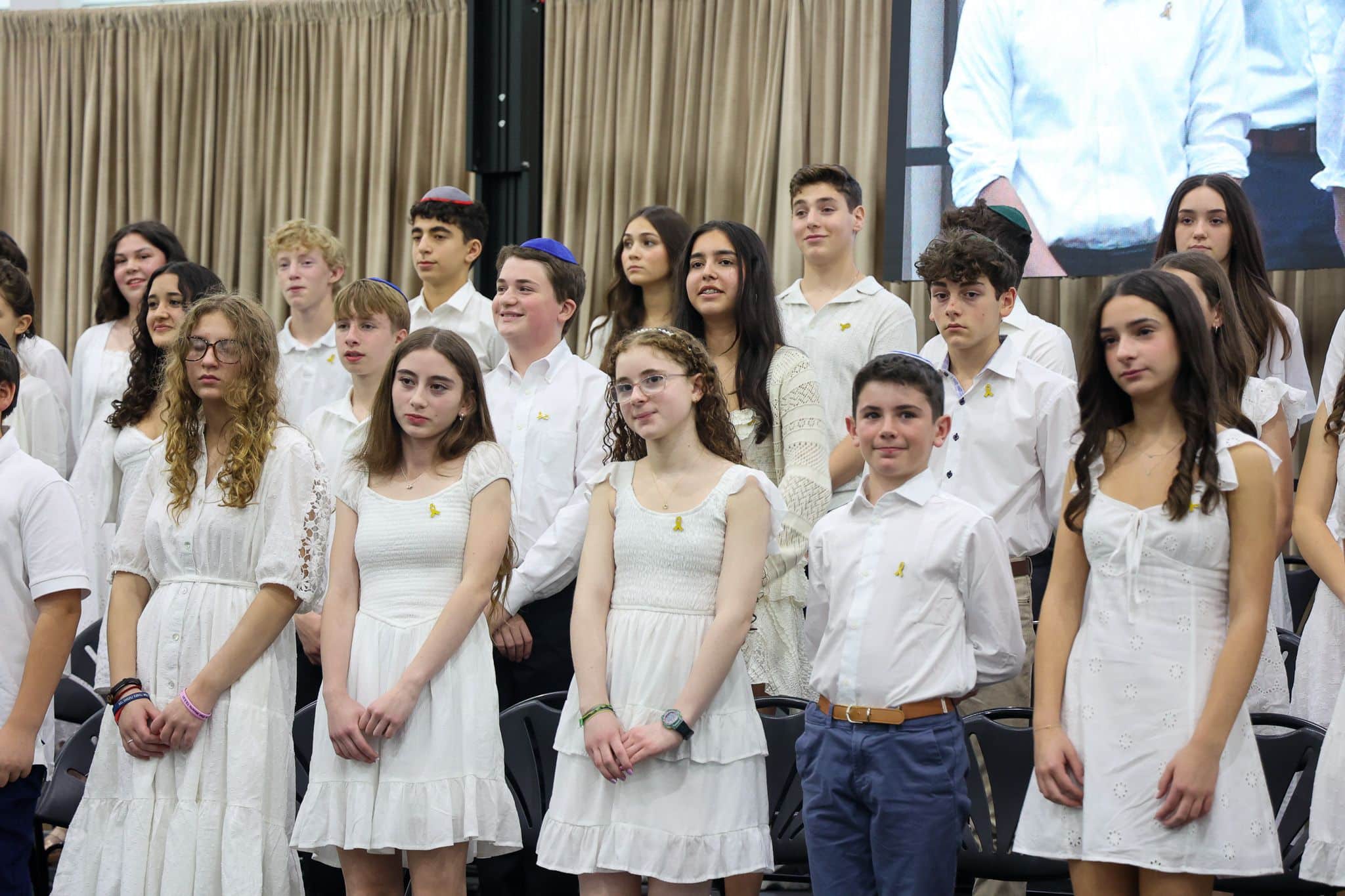 A group of young people stands indoors, wearing white outfits. They appear attentive and composed, with some looking toward a screen in the background.