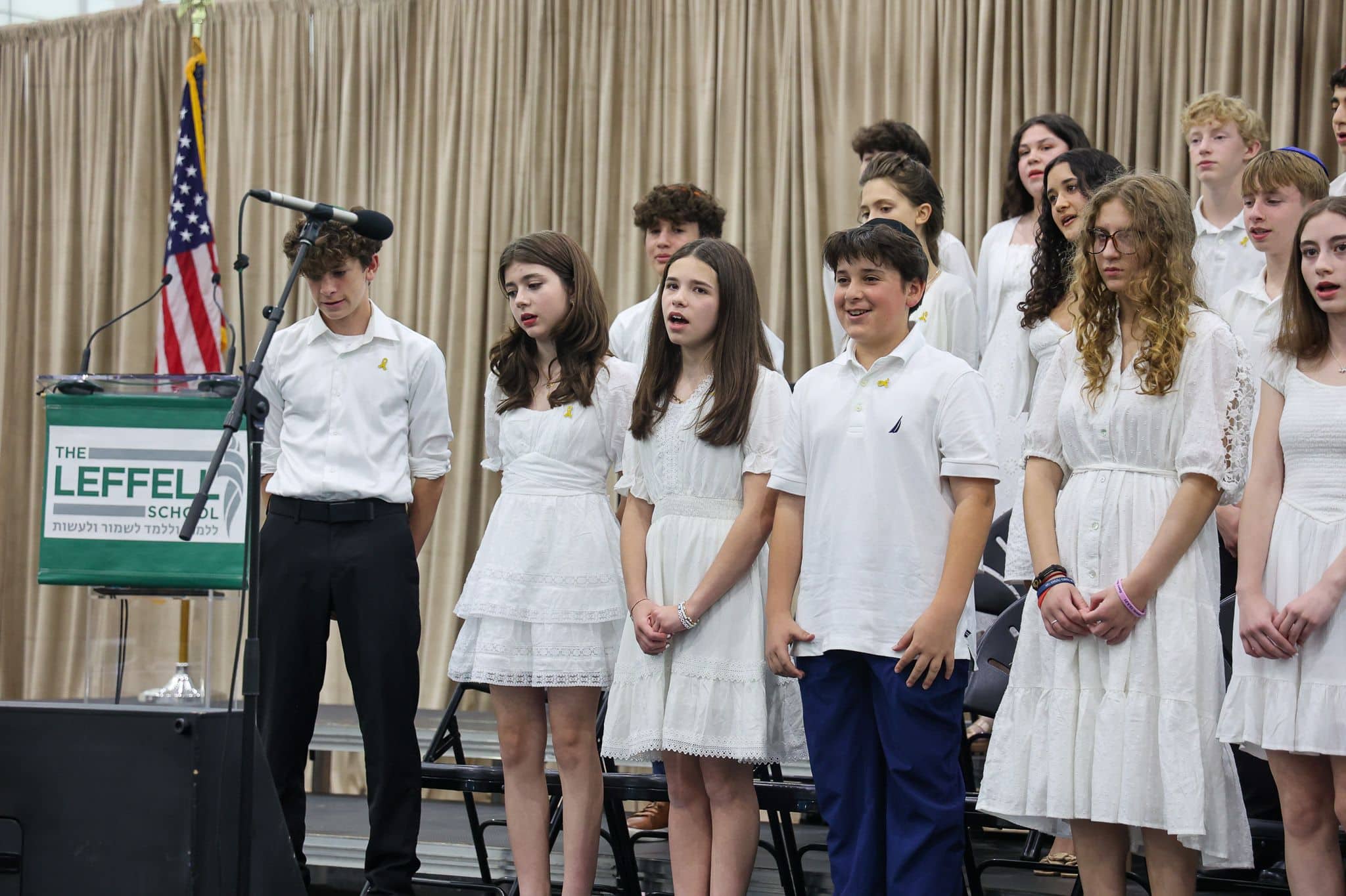 A group of young people stands indoors, wearing white outfits. They appear attentive and composed, with some looking toward a screen in the background.