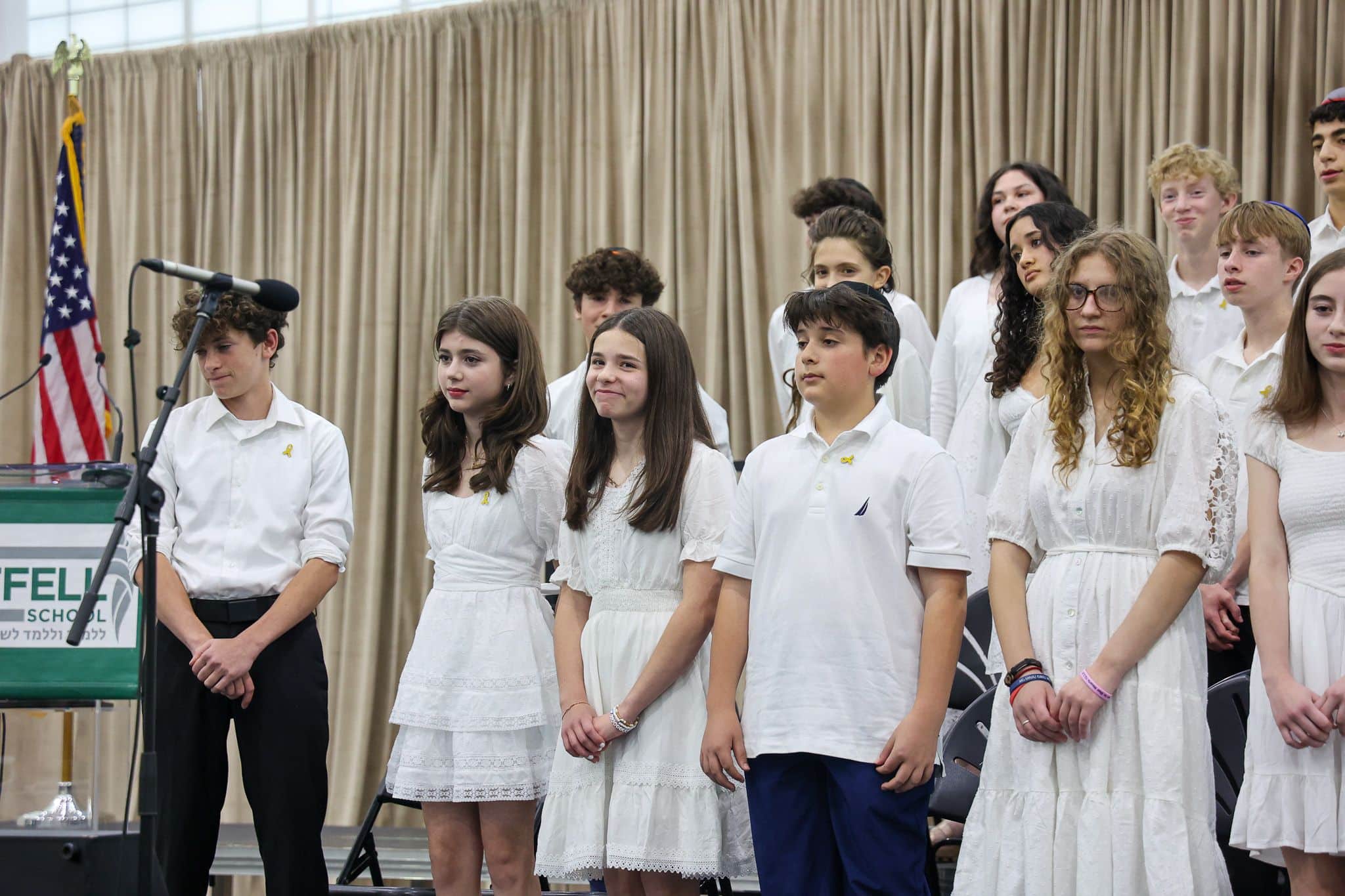 A group of young people stands indoors, wearing white outfits. They appear attentive and composed, with some looking toward a screen in the background.