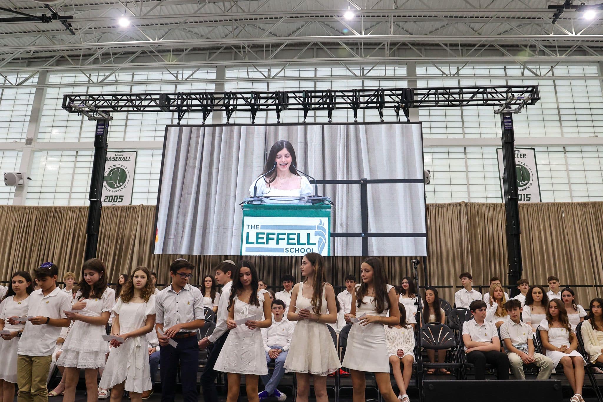 A young person speaks at a podium labeled "The Leffell School" on a large screen. Seated in front, students in white attire listen attentively. The setting is academic and formal, with a backdrop of stage curtains and an American flag.