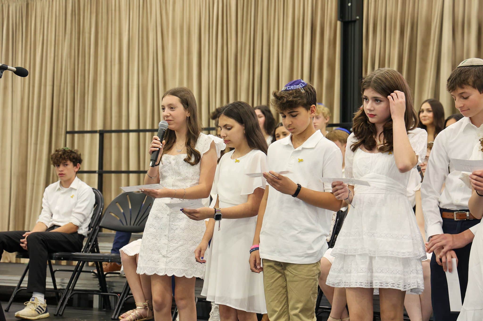 A group of young people stands indoors, wearing white outfits. They appear attentive and composed, with some looking toward a screen in the background.