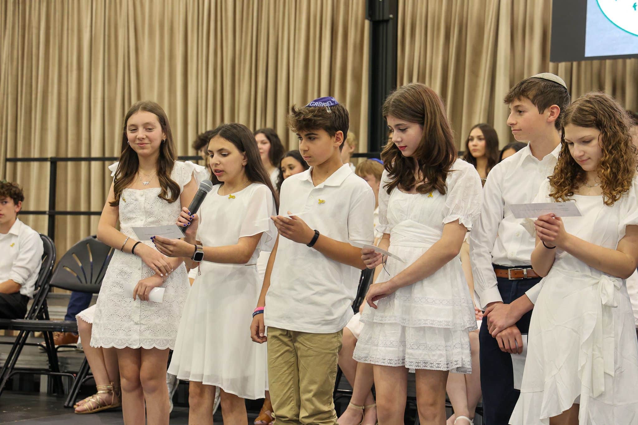 A group of young people stands indoors, wearing white outfits. They appear attentive and composed, with some looking toward a screen in the background.