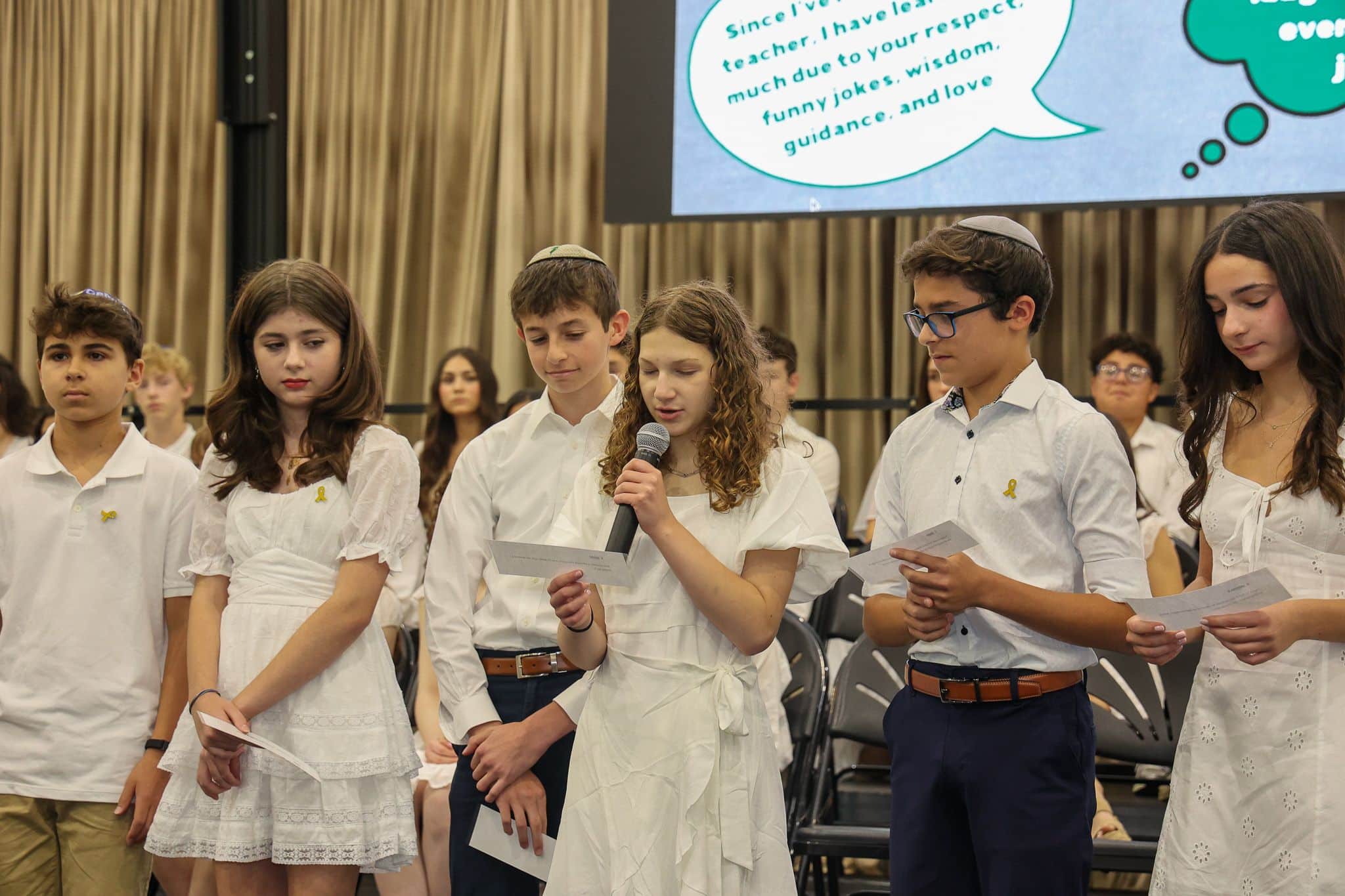 A group of young students stands in a line, dressed in formal white attire. A girl speaks into a microphone while the others hold papers, conveying a sense of ceremony. A large screen in the background shows a message of appreciation, creating an atmosphere of reflection and gratitude.