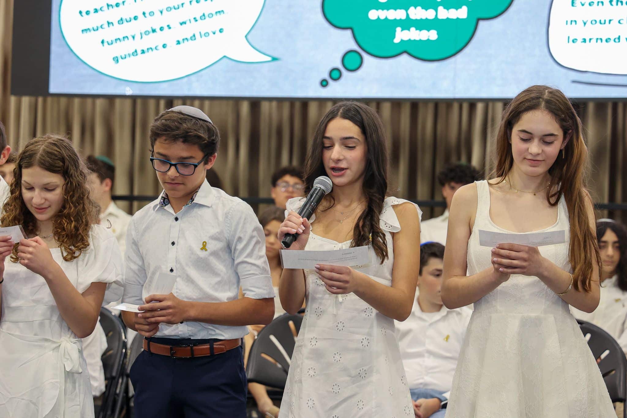 A group of young students stands in a line, dressed in formal white attire. A girl speaks into a microphone while the others hold papers, conveying a sense of ceremony. A large screen in the background shows a message of appreciation, creating an atmosphere of reflection and gratitude.