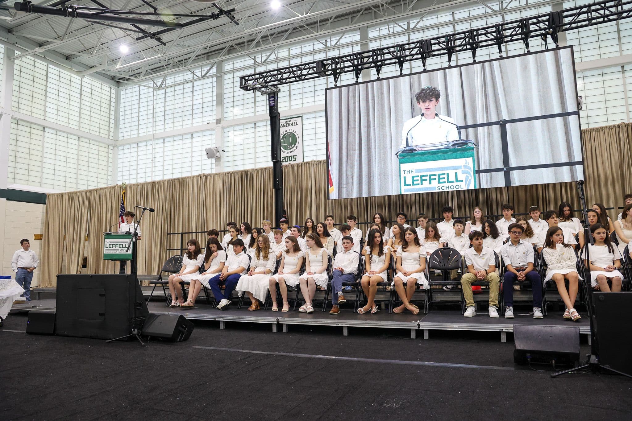 A young person speaks at a podium labeled "The Leffell School" on a large screen. Seated in front, students in white attire listen attentively. The setting is academic and formal, with a backdrop of stage curtains and an American flag.