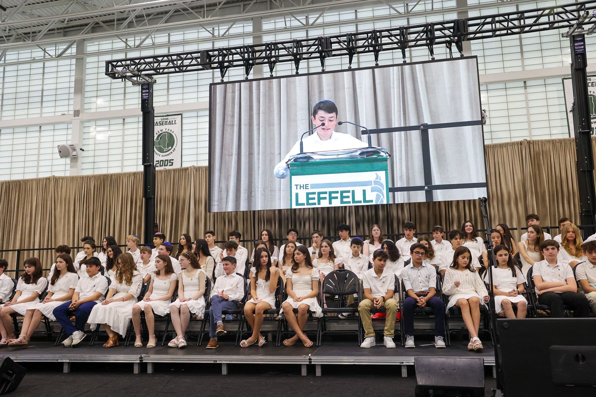 A young person speaks at a podium labeled "The Leffell School" on a large screen. Seated in front, students in white attire listen attentively. The setting is academic and formal, with a backdrop of stage curtains and an American flag.