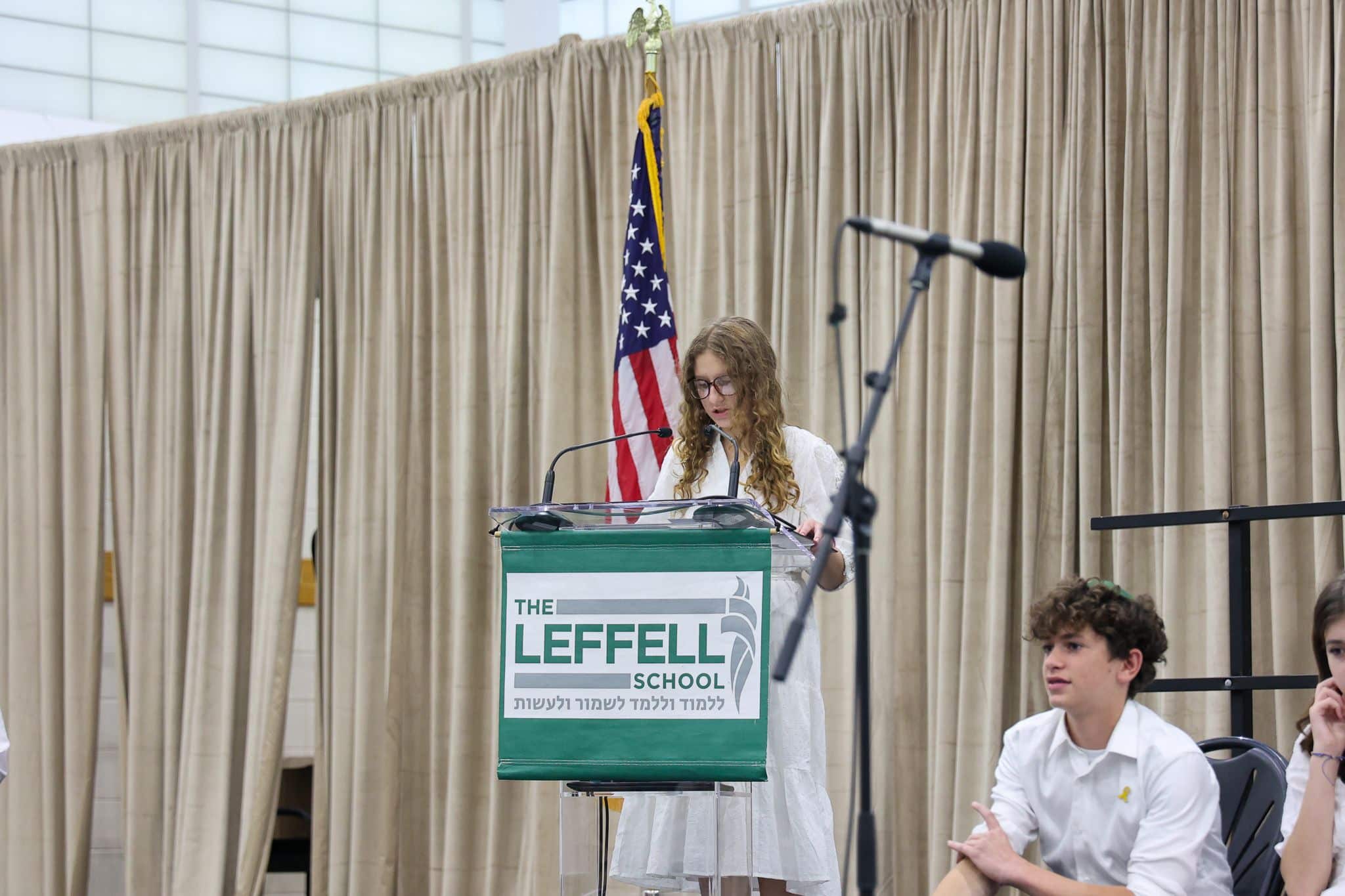 A young person speaks at a podium labeled "The Leffell School," with an American flag on the left. They wear a white outfit, set against beige curtains.
