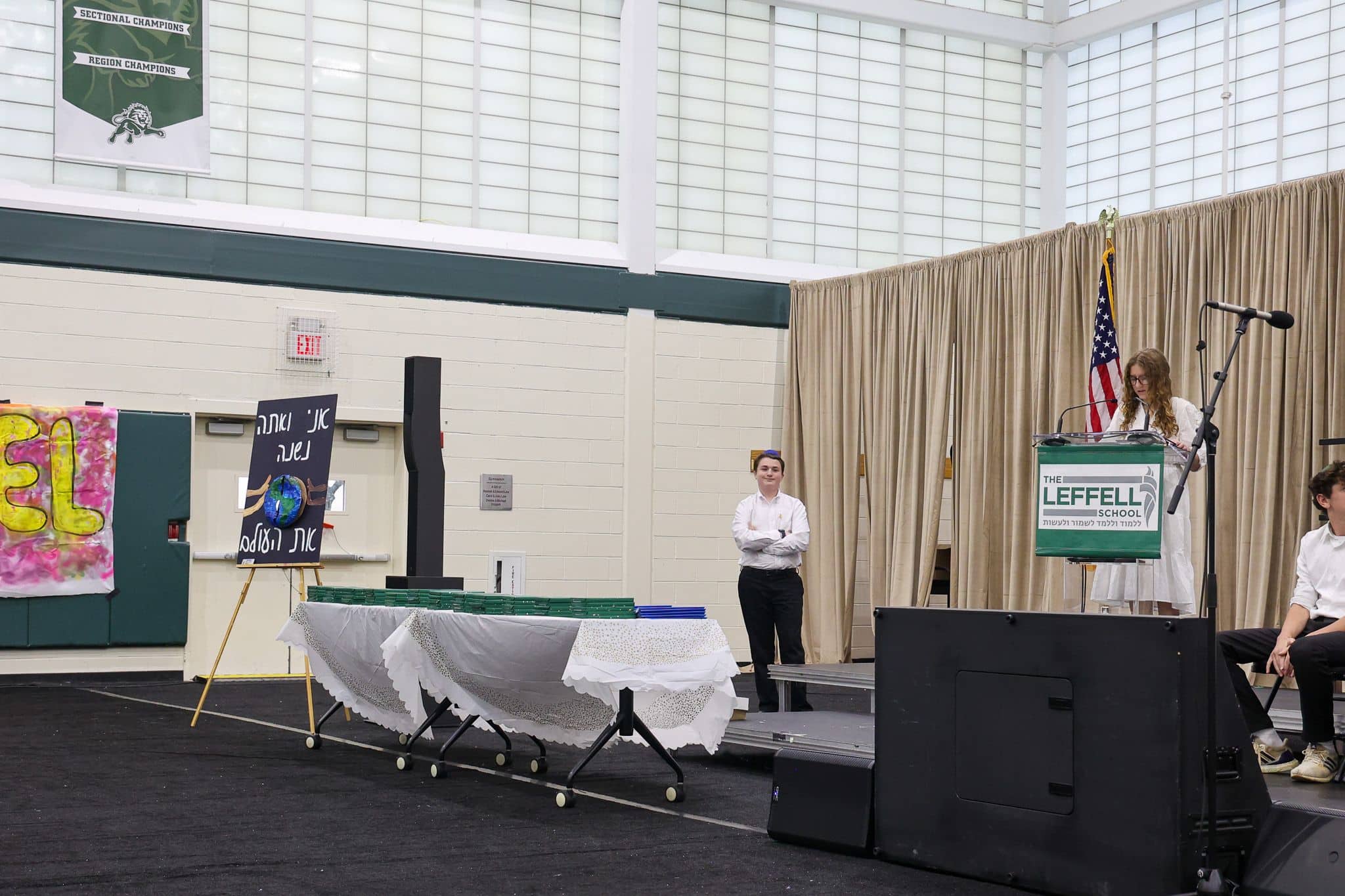 A young person speaks at a podium labeled "The Leffell School," with an American flag on the left. They wear a white outfit, set against beige curtains.
