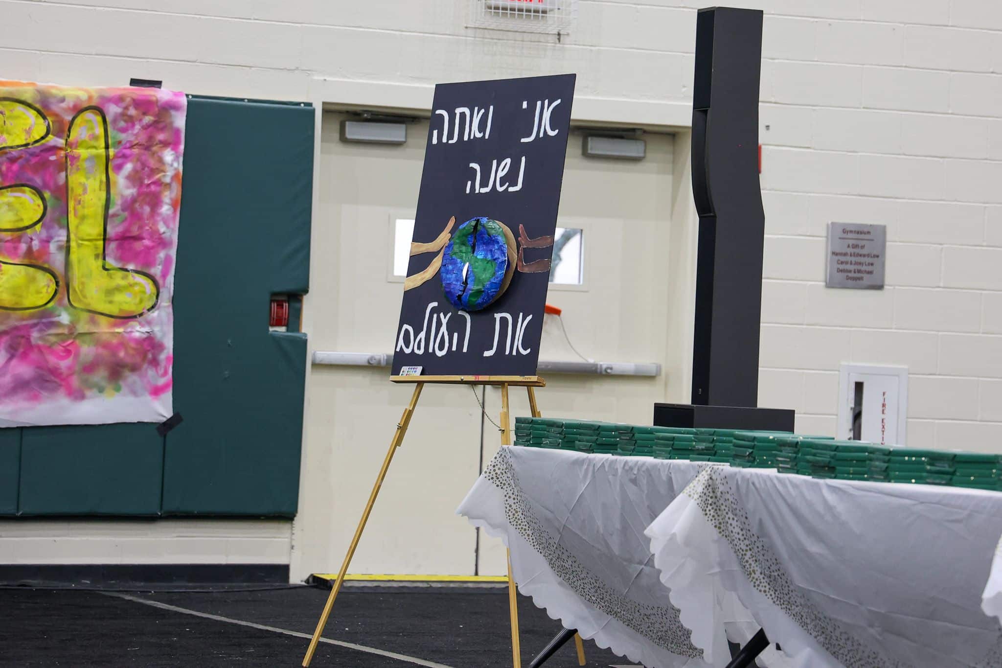 A decorated easel displays a Earth-themed artwork with Hebrew text in a gym or hall. Nearby, a colorful banner and tables with green items and tablecloths.