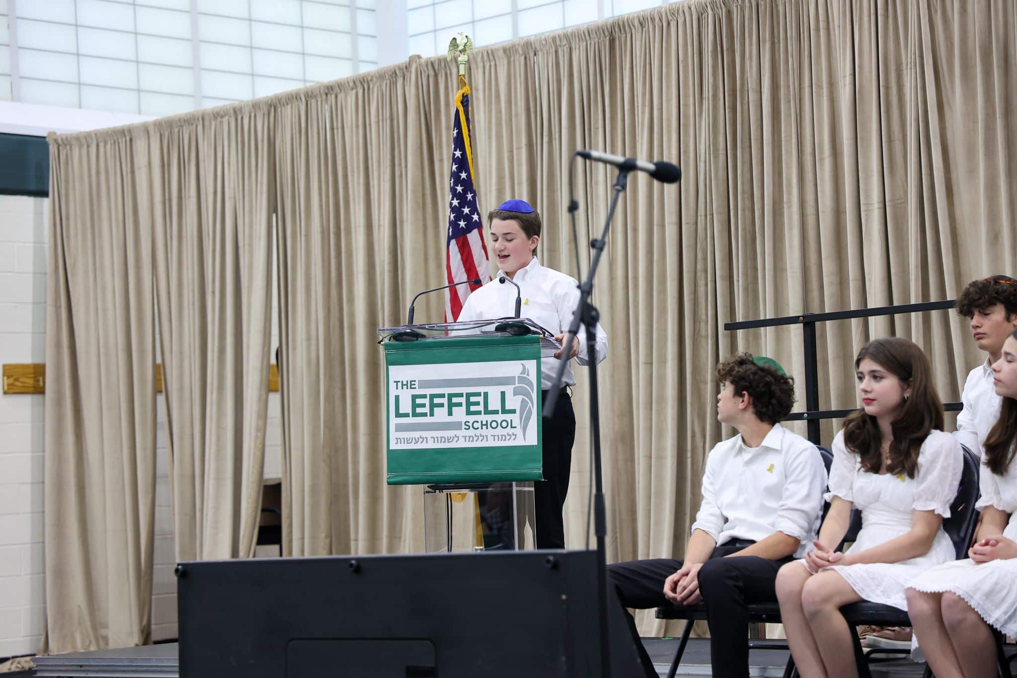 A young person speaks at a podium labeled "The Leffell School," with an American flag on the left. They wear a white outfit, set against beige curtains.