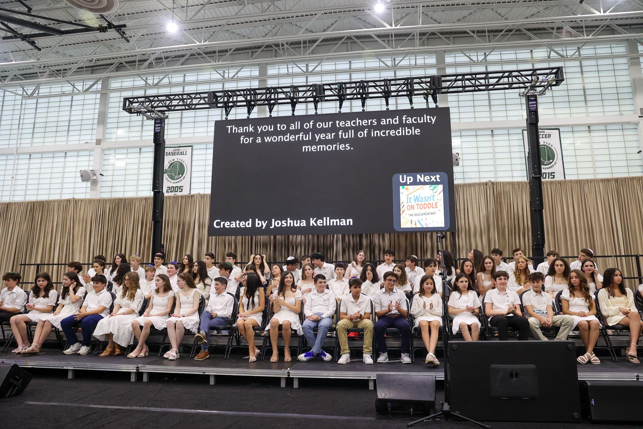 A group of students dressed in white sit on a stage with a large screen behind them displaying the phrase "For certainly we are able" and "Numbers 13:30." A man in a suit stands at a podium to the left, suggesting a formal event, possibly a graduation ceremony. The mood is celebratory and inspirational.