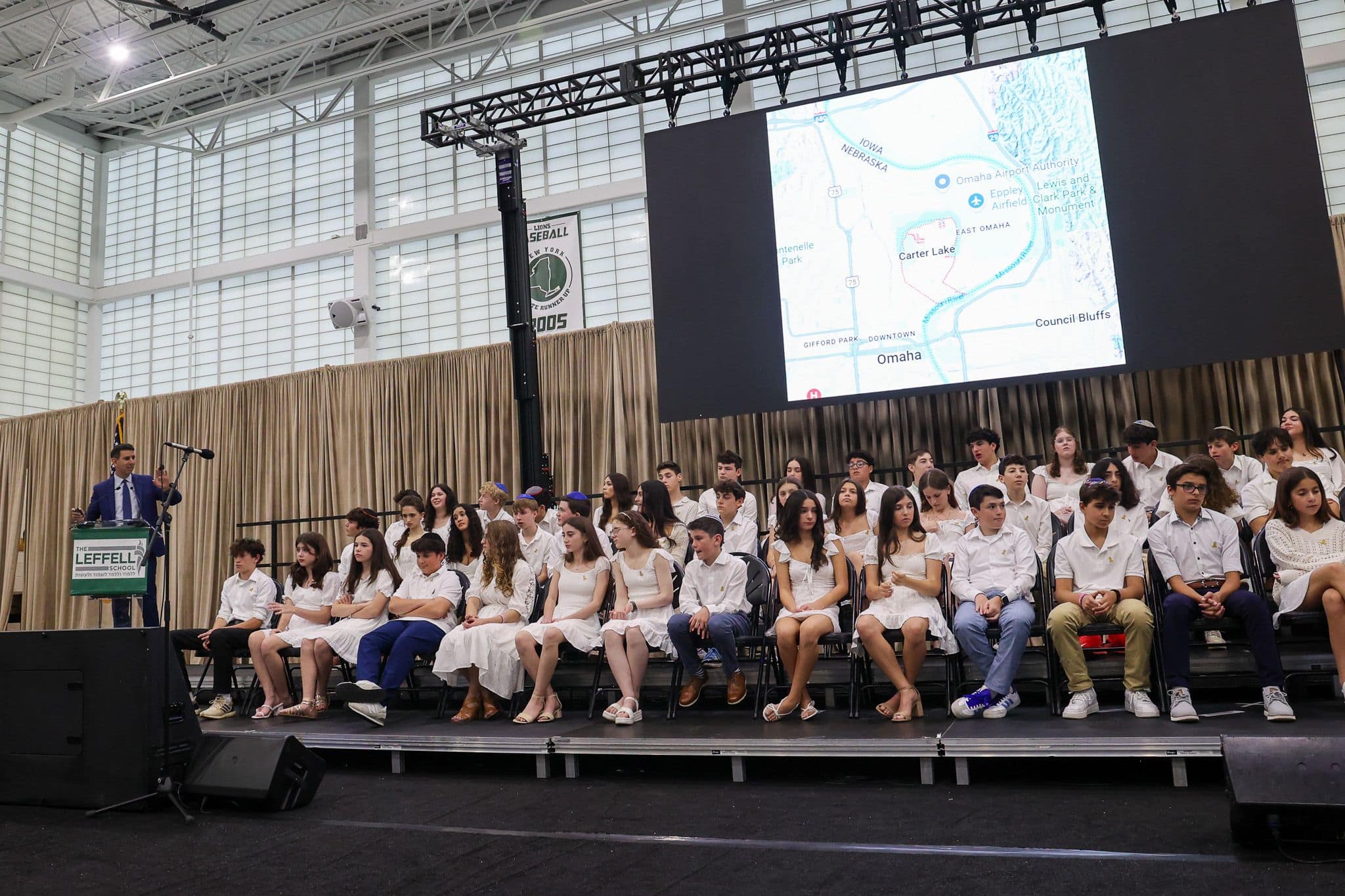 A group of students dressed in white sit on a stage with a large screen behind them displaying the phrase "For certainly we are able" and "Numbers 13:30." A man in a suit stands at a podium to the left, suggesting a formal event, possibly a graduation ceremony. The mood is celebratory and inspirational.