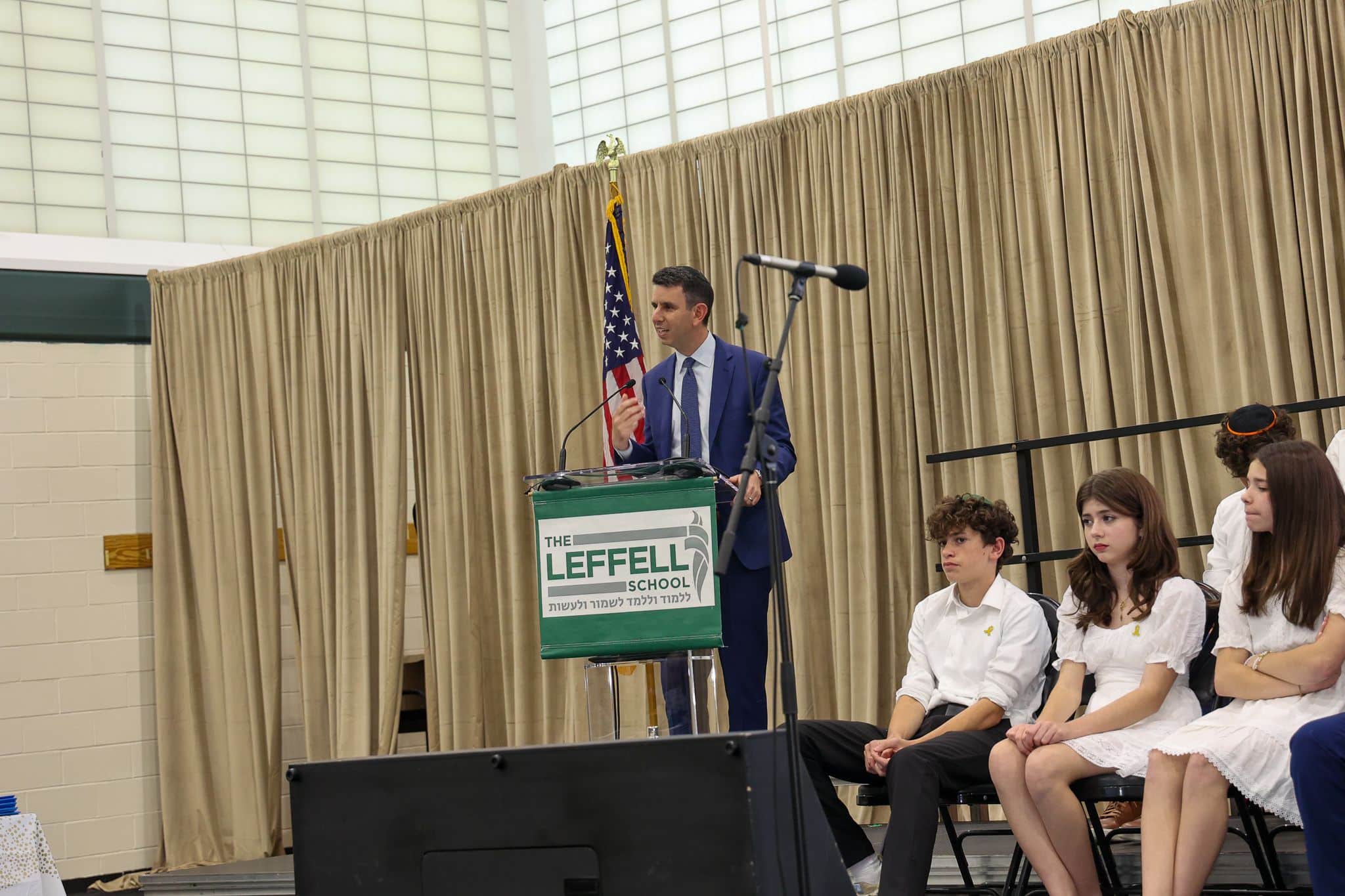 A man in a suit speaks at a podium with "The Leffell School" sign. Behind him is a U.S. flag and beige curtains. Seated are four students in white attire, attentive and serious.