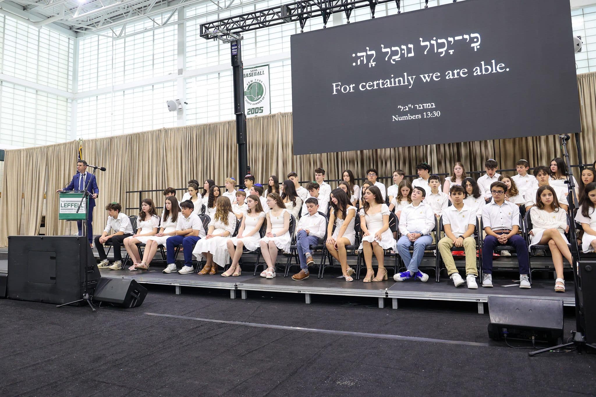 A group of students dressed in white sit on a stage with a large screen behind them displaying the phrase "For certainly we are able" and "Numbers 13:30." A man in a suit stands at a podium to the left, suggesting a formal event, possibly a graduation ceremony. The mood is celebratory and inspirational.