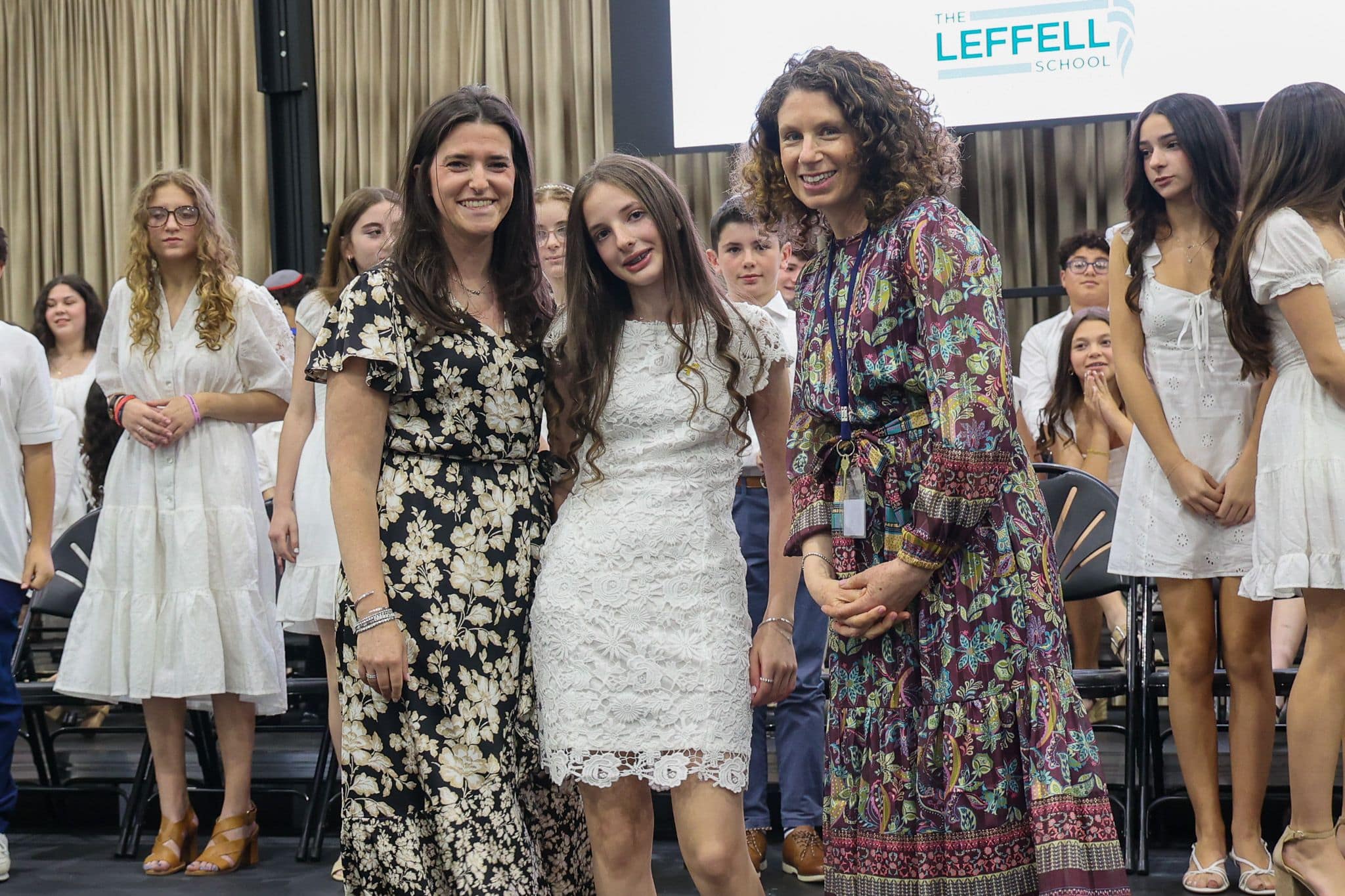 A group of three people, one in the middle and two on each side, are smiling in front of an audience. They are at a ceremony, as shown by the backdrop displaying "The Leffell School." The central figure, a younger girl in a white dress, is flanked by two women in floral dresses. The mood is joyful and celebratory.