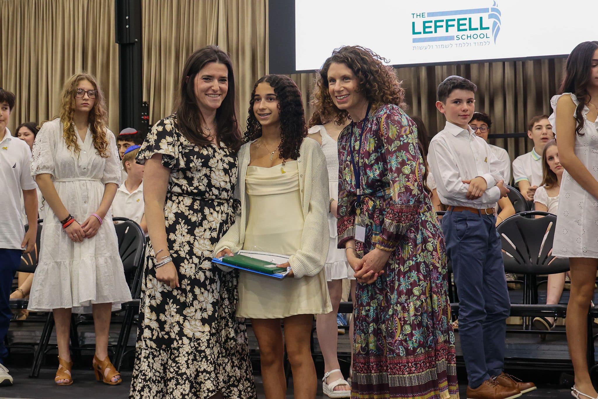 A group of three people, one in the middle and two on each side, are smiling in front of an audience. They are at a ceremony, as shown by the backdrop displaying "The Leffell School." The central figure, a younger girl in a cream dress, is flanked by two women in floral dresses. The mood is joyful and celebratory.
