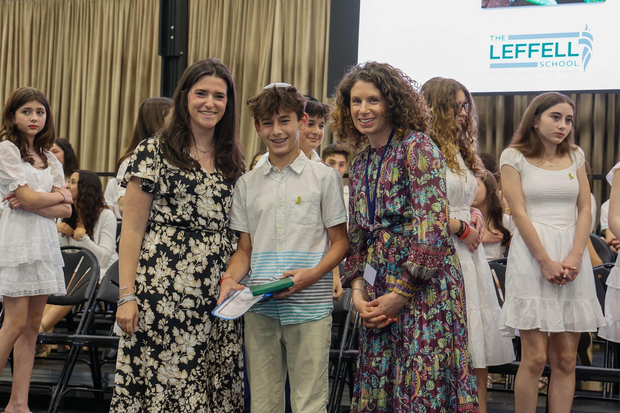 A group of three people, one in the middle and two on each side, are smiling in front of an audience. They are at a ceremony, as shown by the backdrop displaying "The Leffell School." The central figure, a younger boy in a white top, is flanked by two women in floral dresses. The mood is joyful and celebratory.