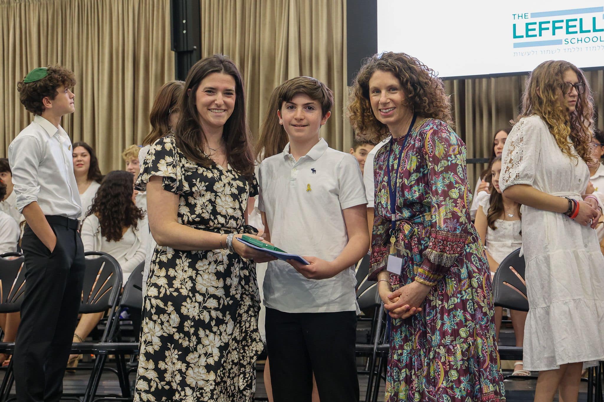 A group of three people, one in the middle and two on each side, are smiling in front of an audience. They are at a ceremony, as shown by the backdrop displaying "The Leffell School." The central figure, a younger boy in a white top, is flanked by two women in floral dresses. The mood is joyful and celebratory.