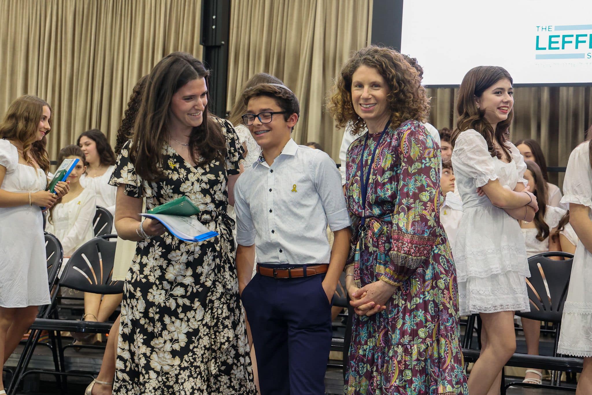 A group of three people, one in the middle and two on each side, are smiling in front of an audience. They are at a ceremony, as shown by the backdrop displaying "The Leffell School." The central figure, a younger boy in a white top, is flanked by two women in floral dresses. The mood is joyful and celebratory.