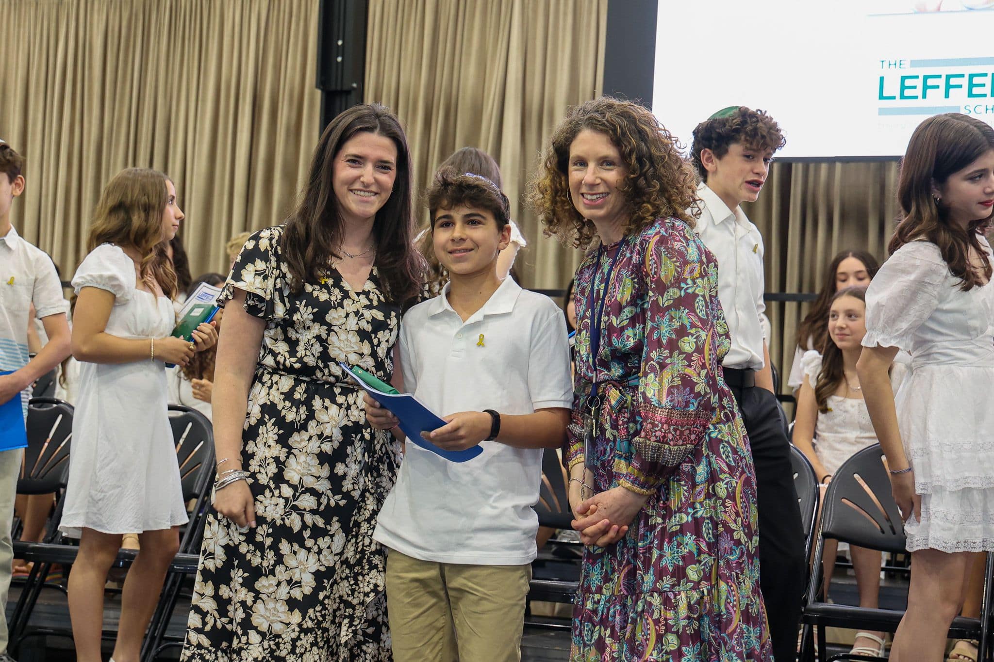 A group of three people, one in the middle and two on each side, are smiling for camera. They are at a ceremony, as shown by the backdrop displaying "The Leffell School." The central figure, a younger student in white clothes , on the sides are two women in floral dresses.