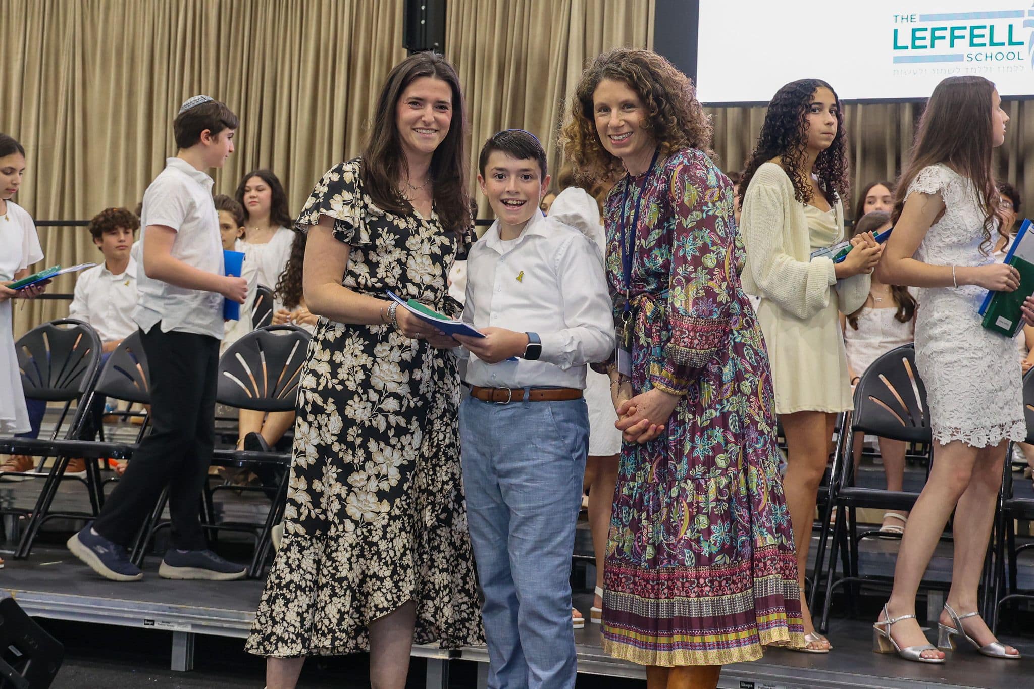 A group of three people, one in the middle and two on each side, are smiling for camera. They are at a ceremony, as shown by the backdrop displaying "The Leffell School." The central figure, a younger student in white clothes , on the sides are two women in floral dresses.