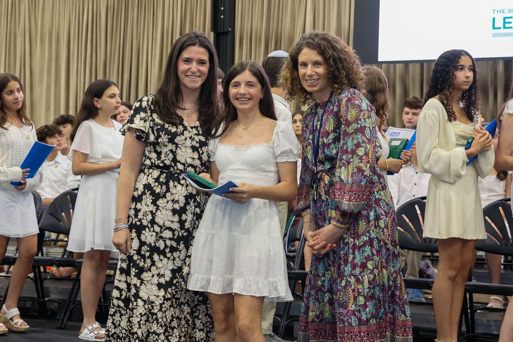 A group of three people, one in the middle and two on each side, are smiling for camera. They are at a ceremony, as shown by the backdrop displaying "The Leffell School." The central figure, a younger student in white clothes , on the sides are two women in floral dresses.