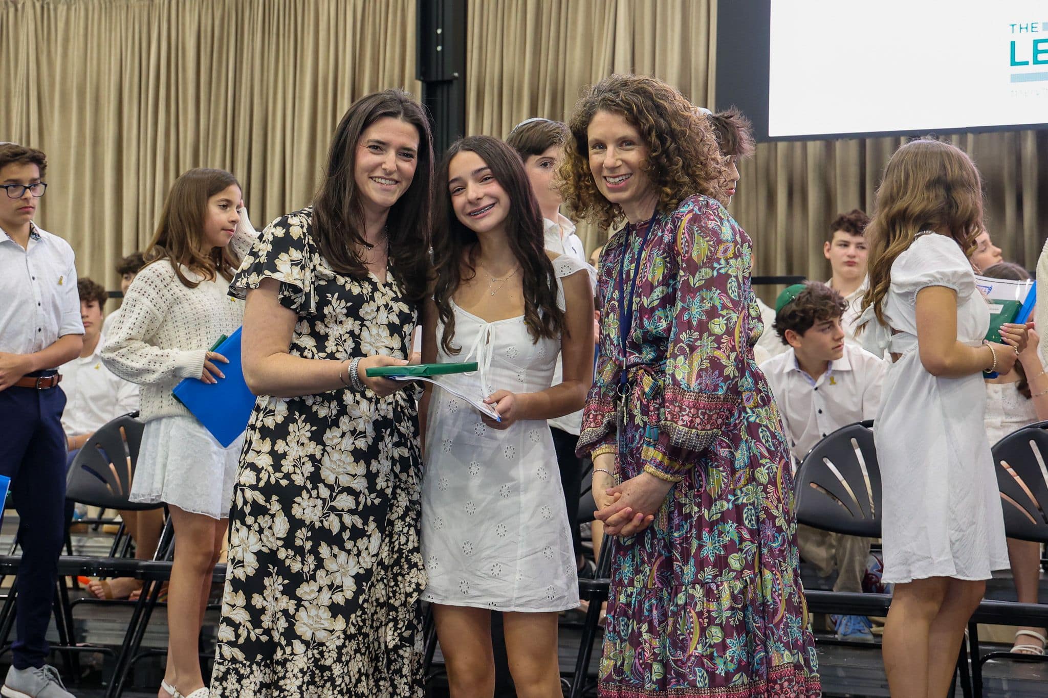 A group of three people, one in the middle and two on each side, are smiling for camera. They are at a ceremony, as shown by the backdrop displaying "The Leffell School." The central figure, a younger student in white clothes , on the sides are two women in floral dresses.