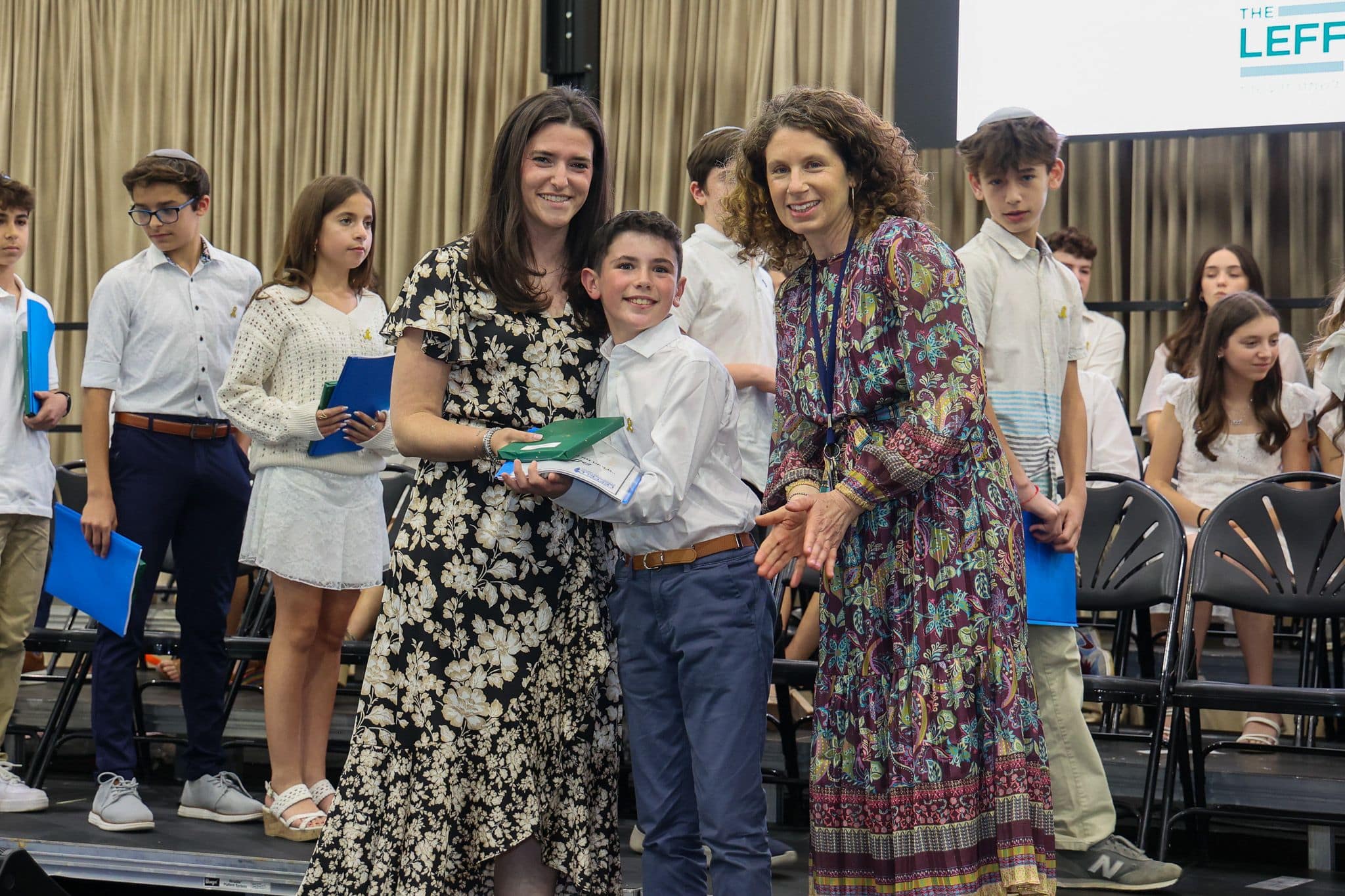 A group of three people, one in the middle and two on each side, are smiling for camera. They are at a ceremony, as shown by the backdrop displaying "The Leffell School." The central figure, a younger student in white clothes , on the sides are two women in floral dresses.