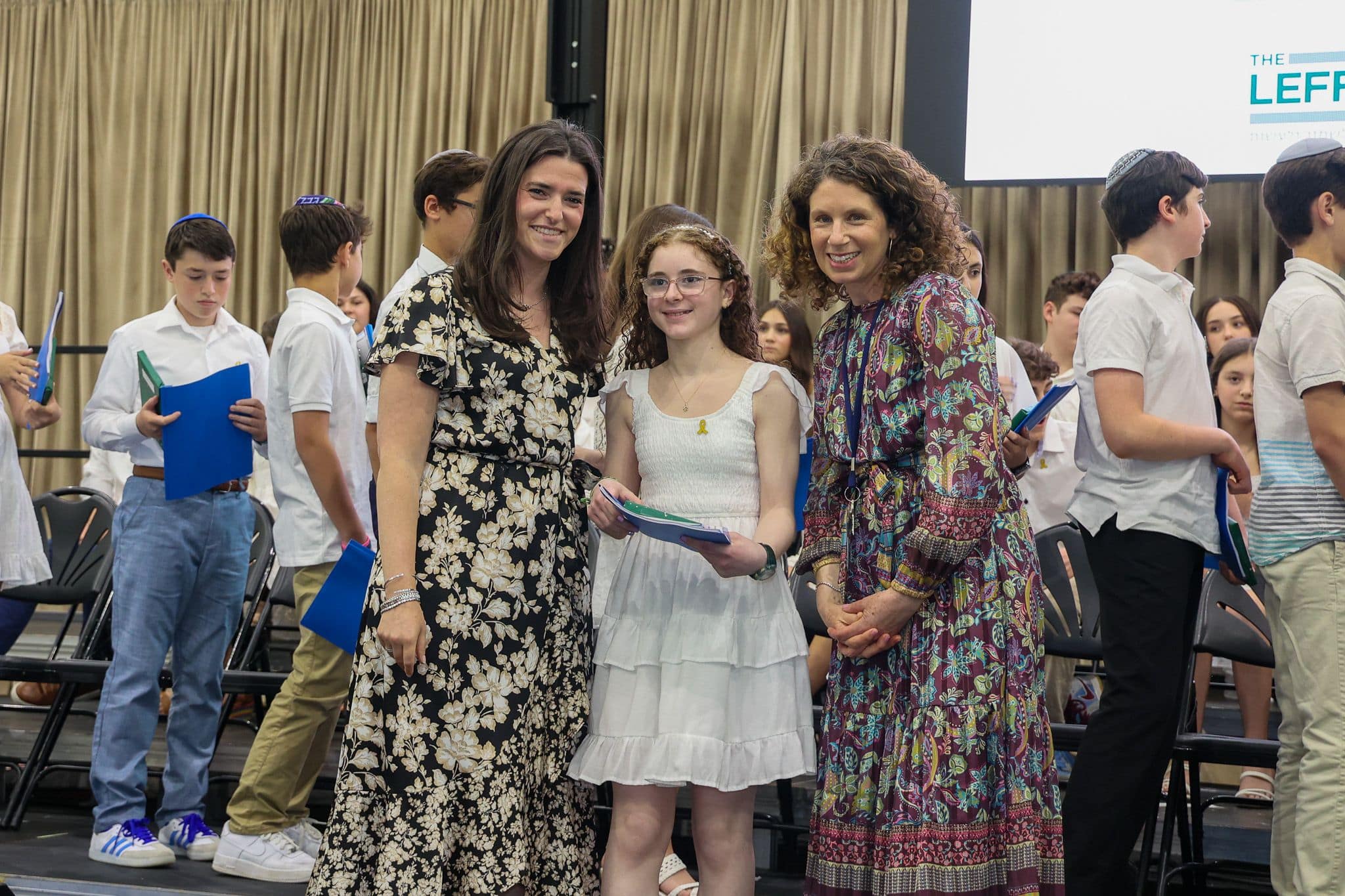 A group of three people, one in the middle and two on each side, are smiling for camera. They are at a ceremony, as shown by the backdrop displaying "The Leffell School." The central figure, a younger student in white clothes , on the sides are two women in floral dresses.