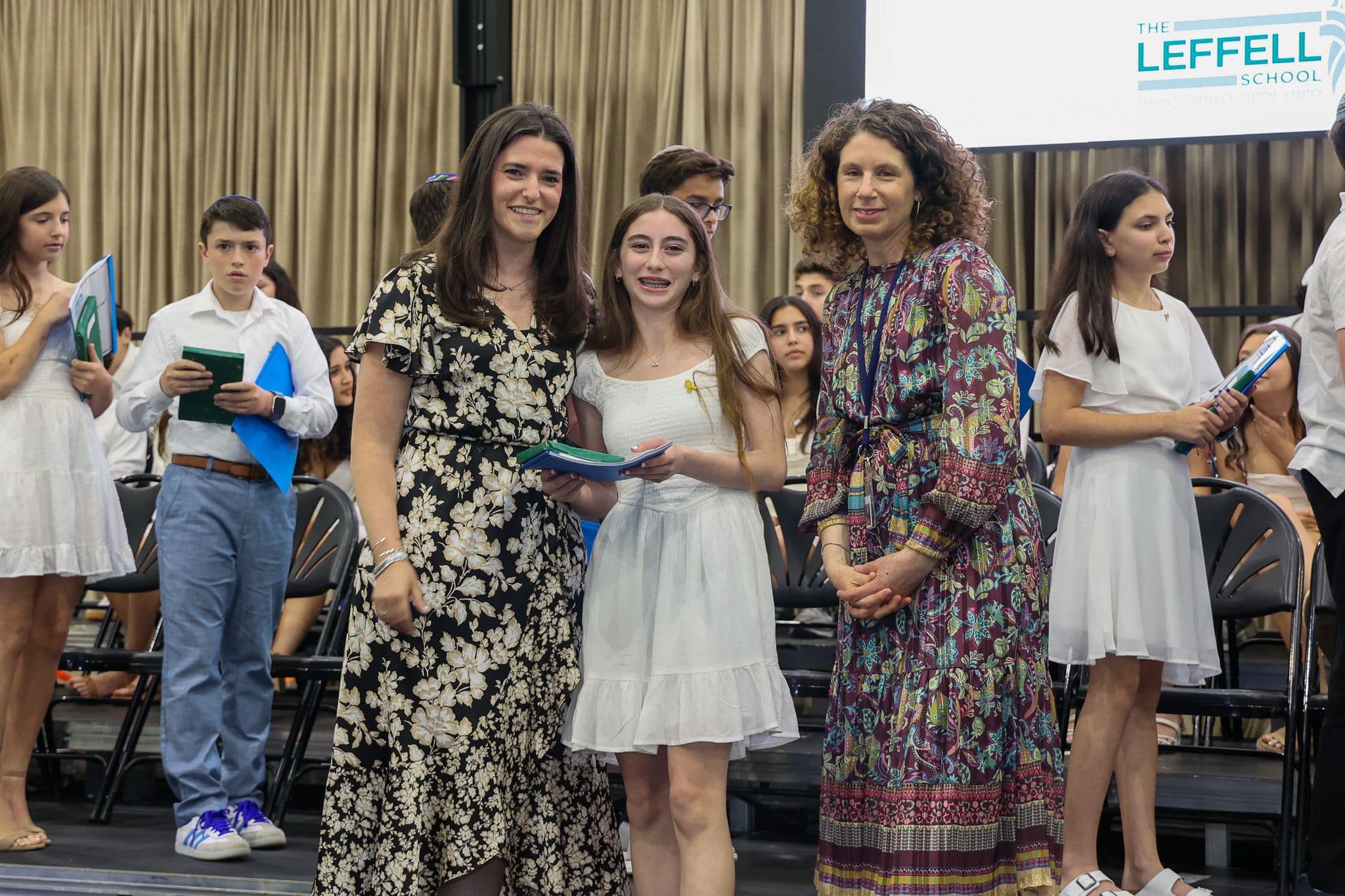 A group of three people, one in the middle and two on each side, are smiling for camera. They are at a ceremony, as shown by the backdrop displaying "The Leffell School." The central figure, a younger student in white clothes , on the sides are two women in floral dresses.