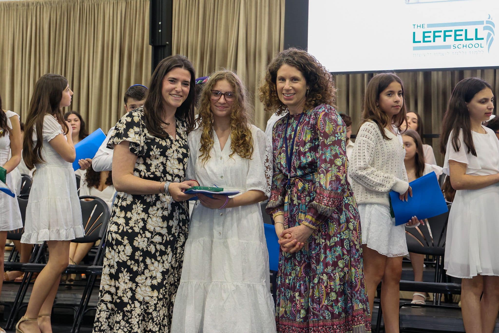 A group of three people, one in the middle and two on each side, are smiling for camera. They are at a ceremony, as shown by the backdrop displaying "The Leffell School." The central figure, a younger student in white clothes , on the sides are two women in floral dresses.