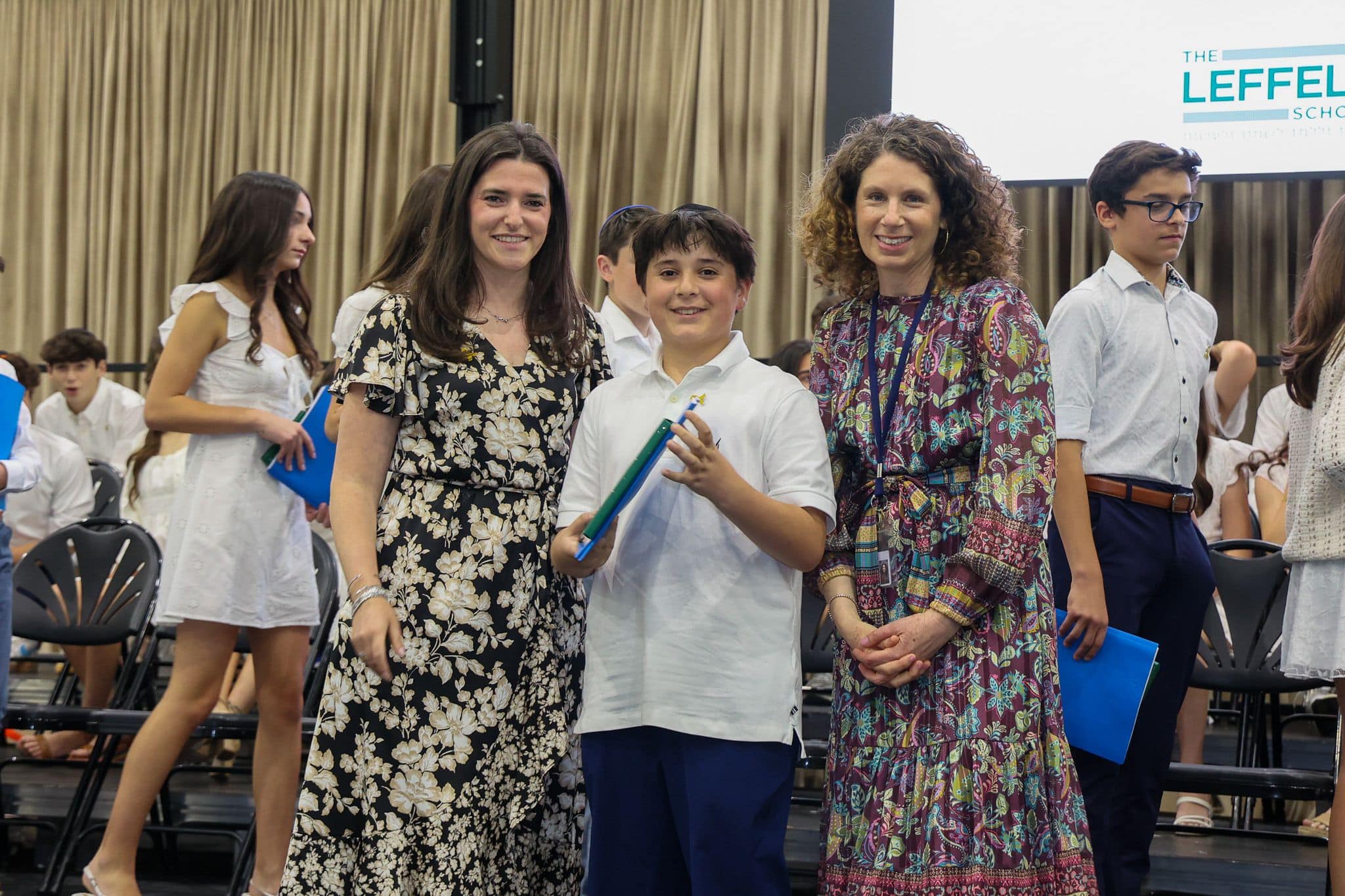 A group of three people, one in the middle and two on each side, are smiling for camera. They are at a ceremony, as shown by the backdrop displaying "The Leffell School." The central figure, a younger student in white clothes , on the sides are two women in floral dresses.