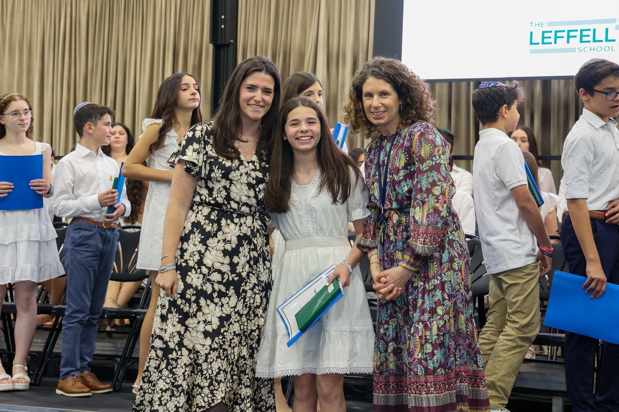 A group of three people, one in the middle and two on each side, are smiling for camera. They are at a ceremony, as shown by the backdrop displaying "The Leffell School." The central figure, a younger student in white clothes , on the sides are two women in floral dresses.