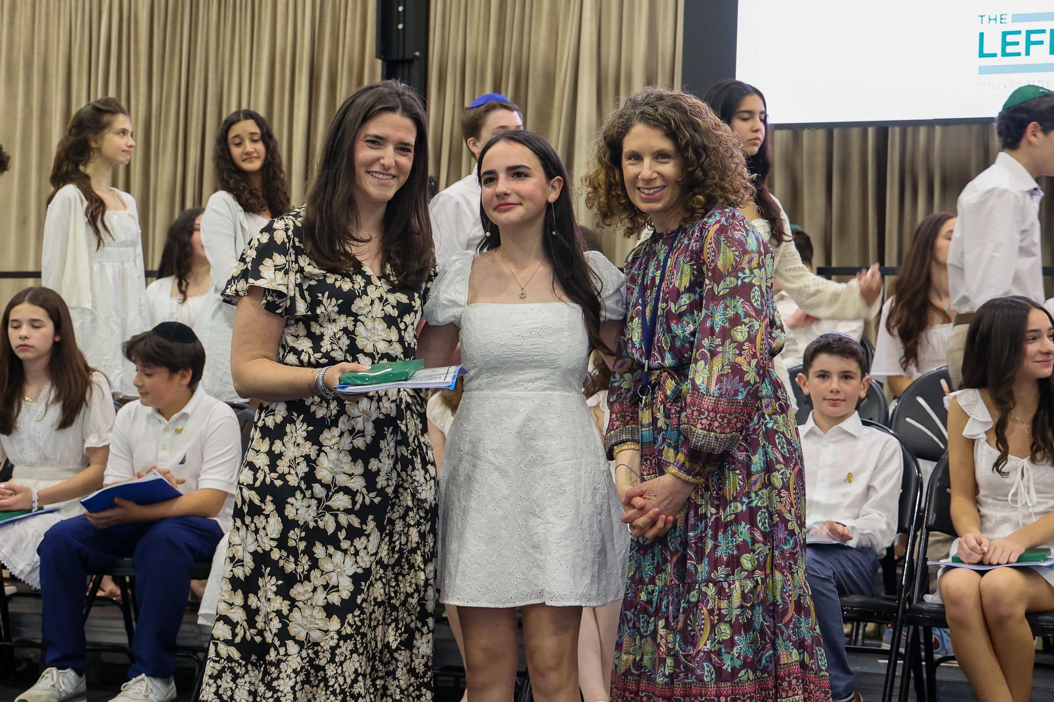 A group of three people, one in the middle and two on each side, are smiling for camera. They are at a ceremony, as shown by the backdrop displaying "The Leffell School." The central figure, a younger student in white clothes , on the sides are two women in floral dresses.