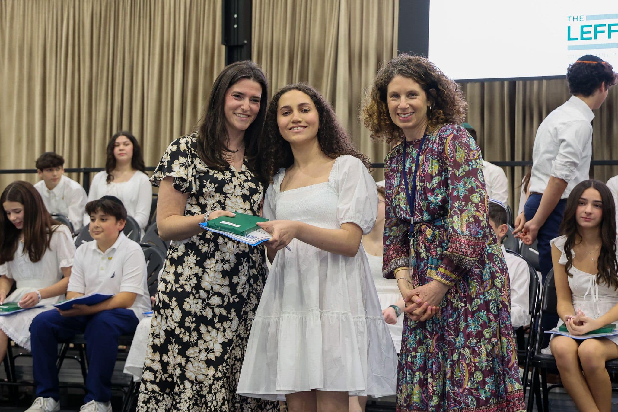 A group of three people, one in the middle and two on each side, are smiling for camera. They are at a ceremony, as shown by the backdrop displaying "The Leffell School." The central figure, a younger student in white clothes , on the sides are two women in floral dresses.