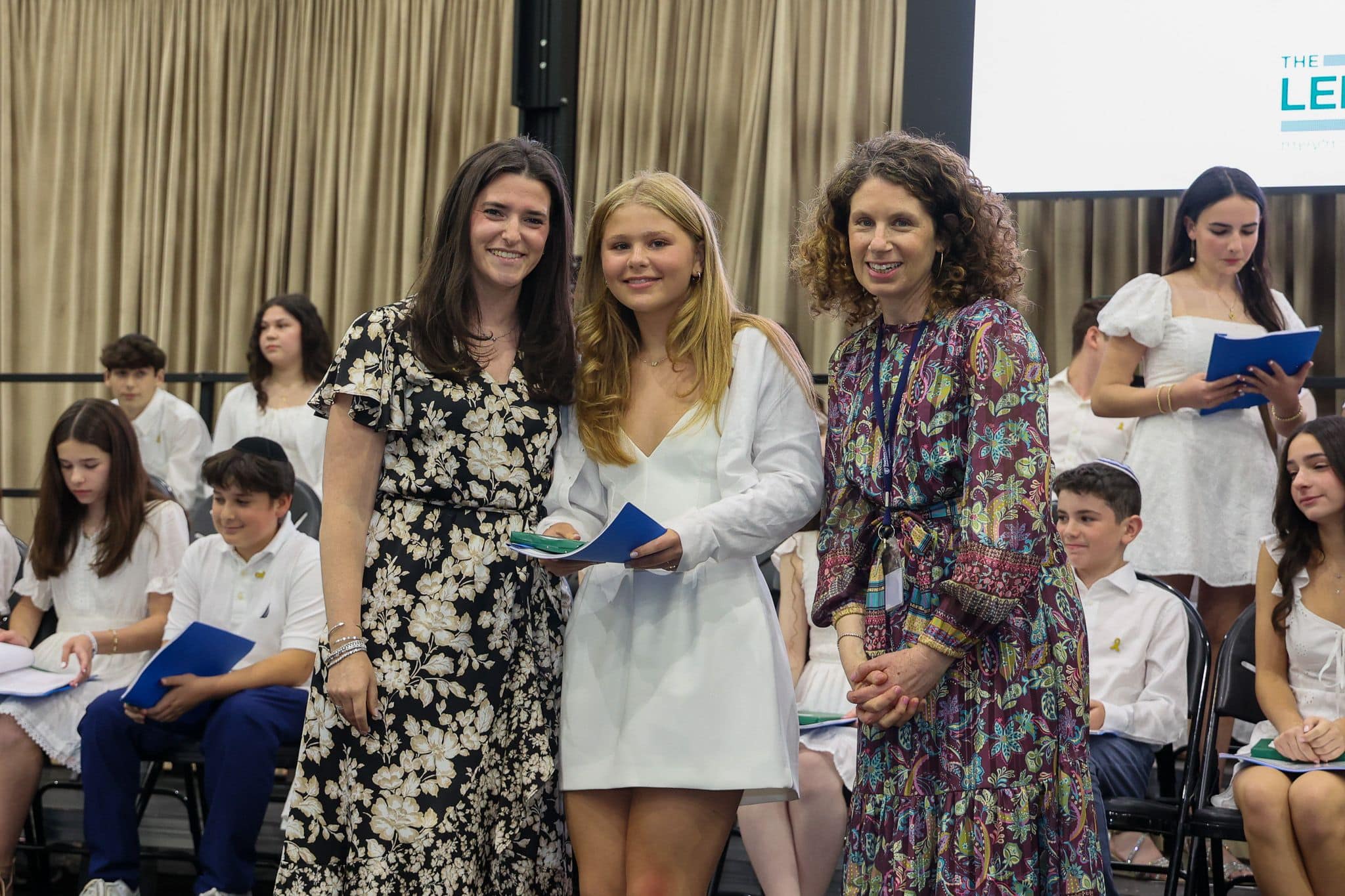 A group of three people, one in the middle and two on each side, are smiling for camera. They are at a ceremony, as shown by the backdrop displaying "The Leffell School." The central figure, a younger student in white clothes , on the sides are two women in floral dresses.