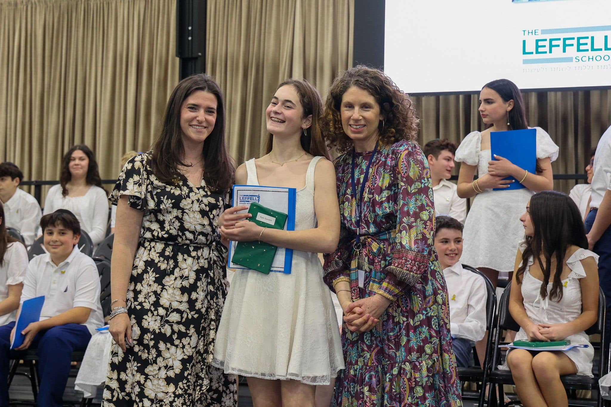 A group of three people, one in the middle and two on each side, are smiling for camera. They are at a ceremony, as shown by the backdrop displaying "The Leffell School." The central figure, a younger student in white clothes , on the sides are two women in floral dresses.